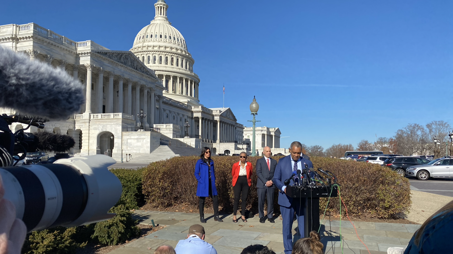 AG Karl Racine addresses reporters from behind a podium in front of the Capitol on a clear day.