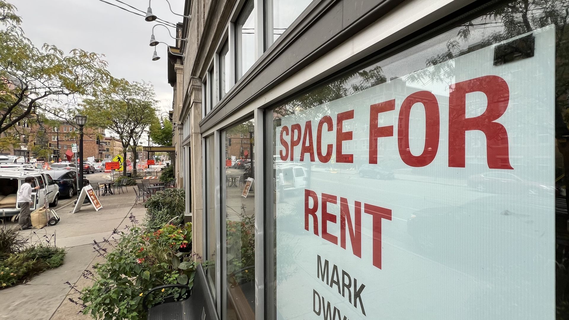 Large red and black "SPACE FOR RENT" sign displayed in the window of a building on a busy urban sidewalk with trees, plants, and cars visible nearby.