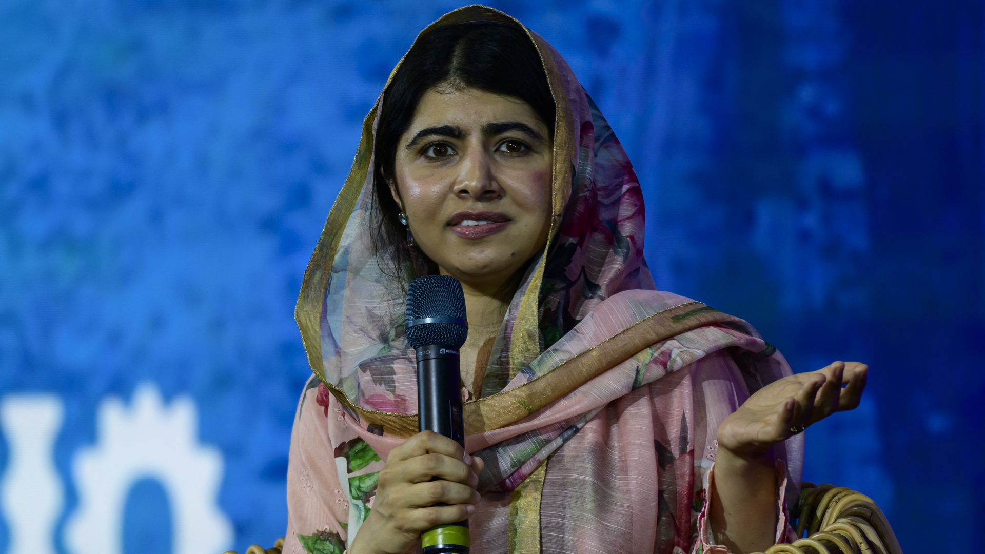 Malala Yousafzai holding a mic and sitting in a seat on a stage.