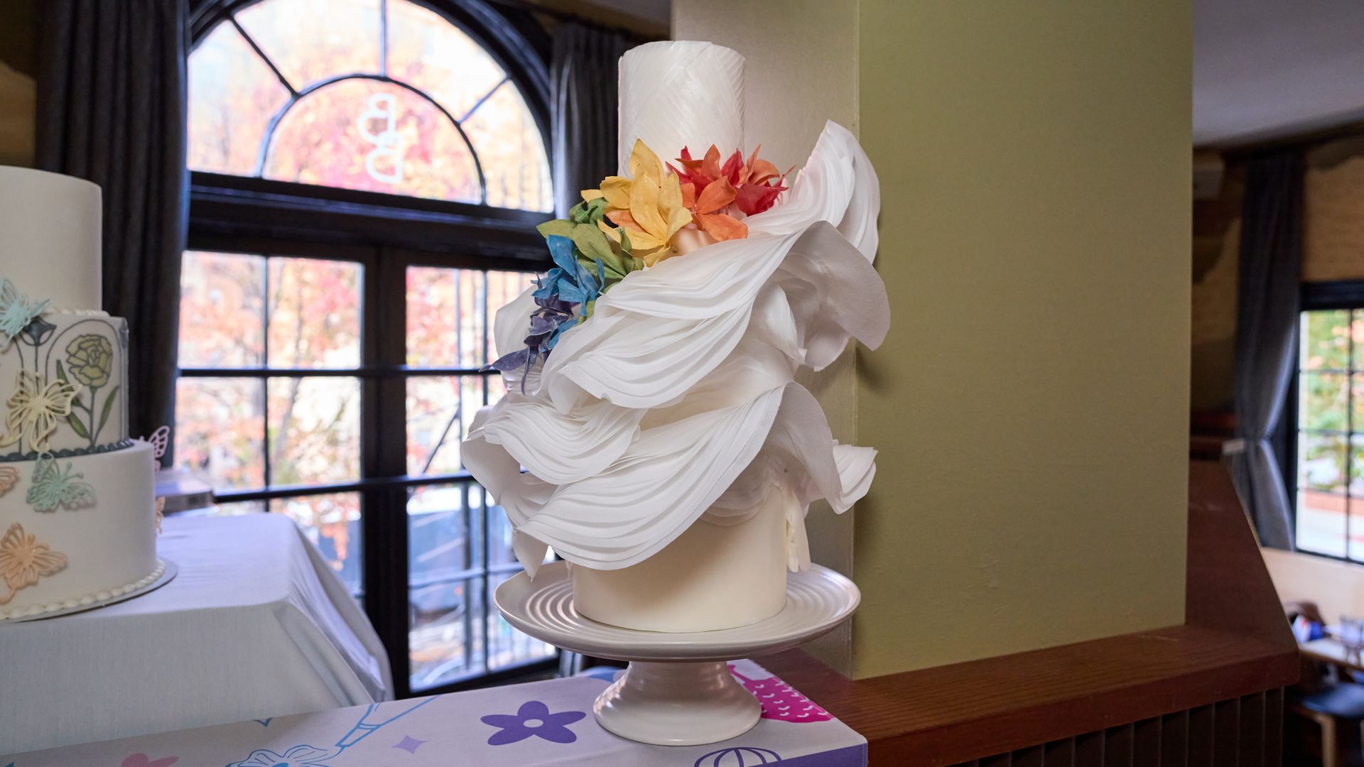 Two cakes on a table in a room with an arched window: a white tiered cake with pastel butterfly decorations, and a draped column topped with a bright rainbow flower arrangement.