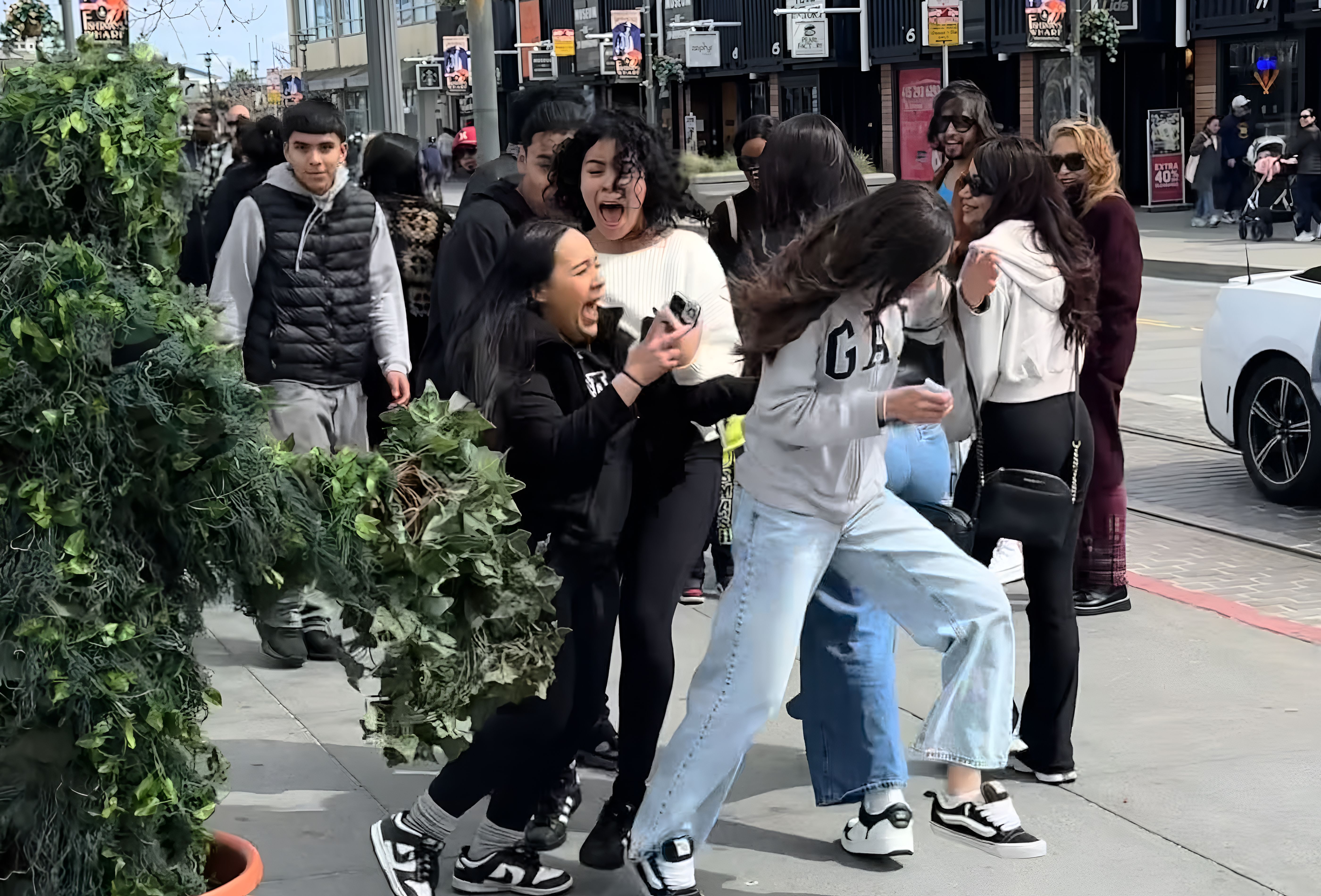 Group of people on a city street reacting with surprise and laughter to a person in leafy green camouflage emerging from a plant pot.