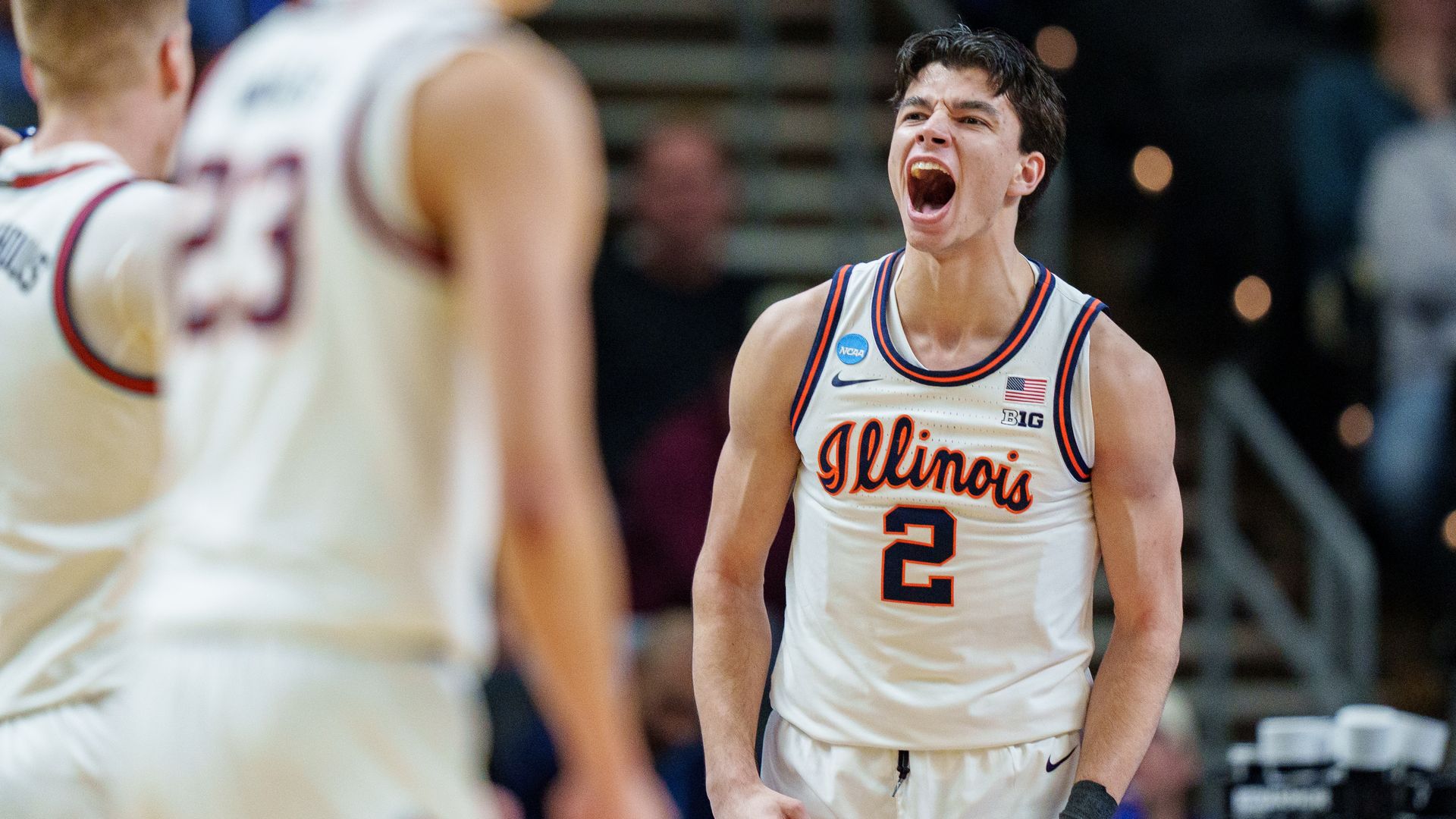 An Illinois basketball player wearing jersey number 2 yells with mouth wide open on the court, while a blurred teammate in white is in the foreground.