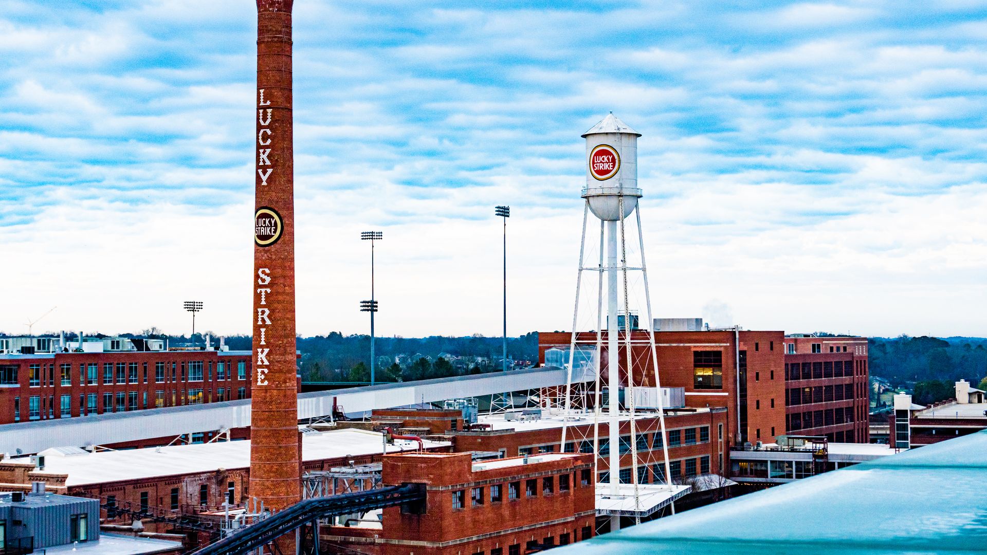 Historic Lucky Strike factory with brick smokestack and white water tower, both branded with the Lucky Strike logo, under a cloudy blue sky.