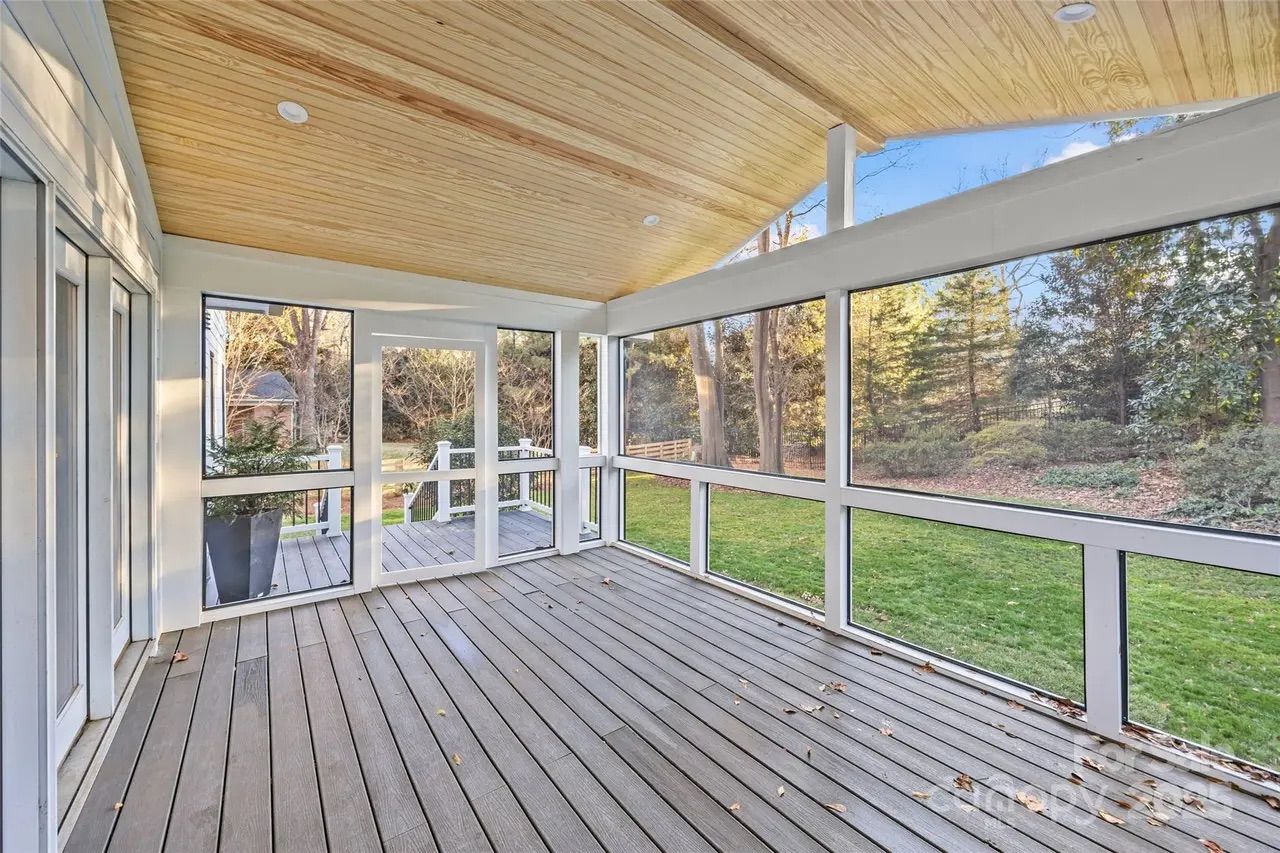 Empty screened porch with wooden deck floor and light wood ceiling, white frame screens, overlooking a green backyard with trees and blue sky.