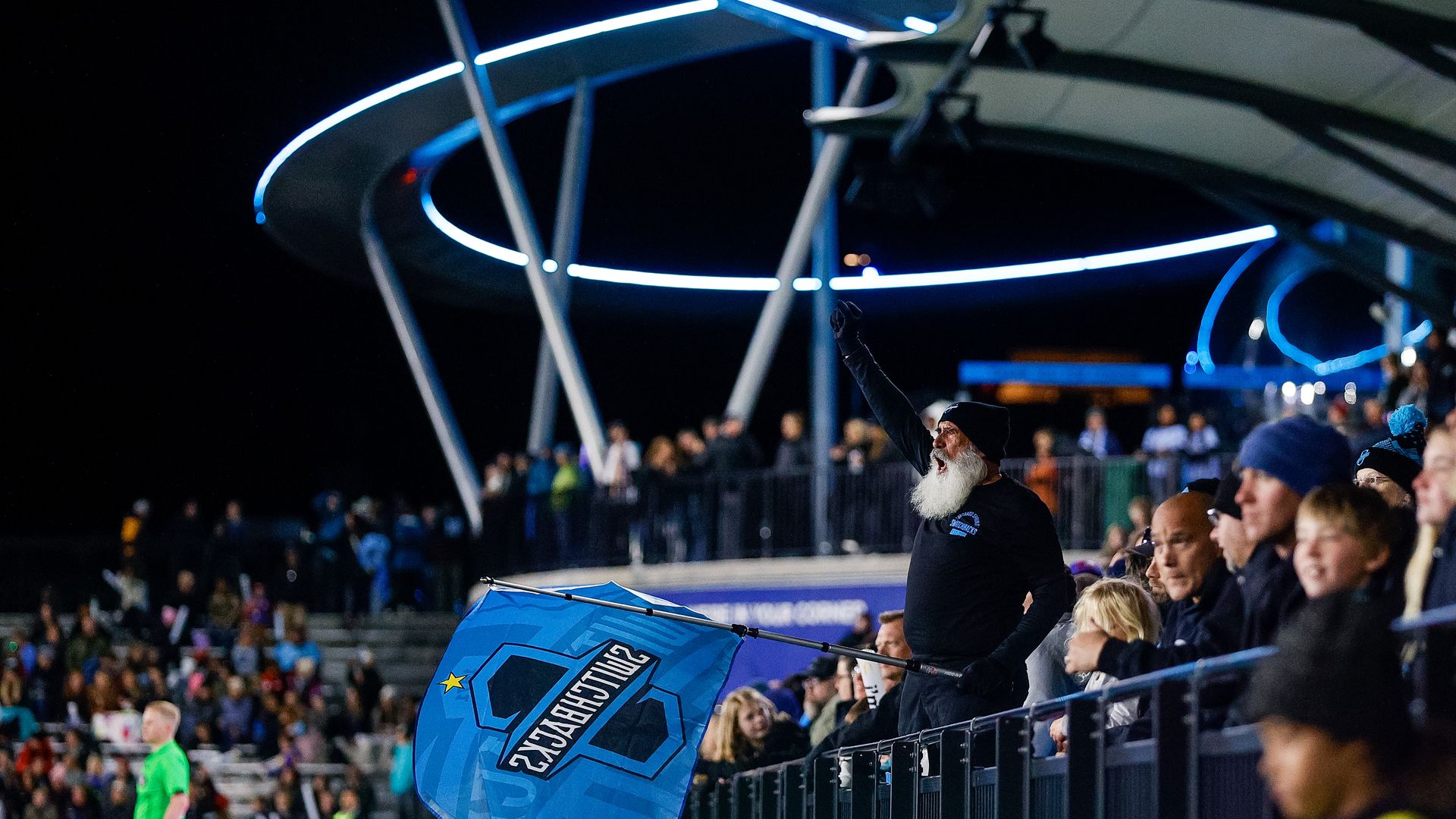 A stadium filled with fans. A man with a white beard, wearing black clothes with the Switchbacks soccer team logo is prominent in the midground, waving a blue Switchbacks flag. Above him is  a swooping steel overhang with blue lighting details.