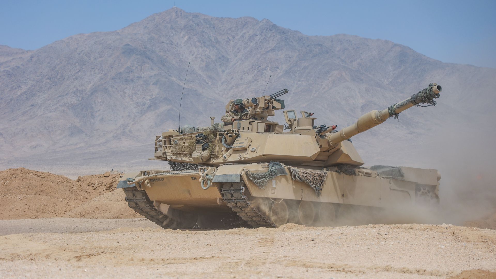 Desert scene with a tan armored tank and a crewman on the turret.