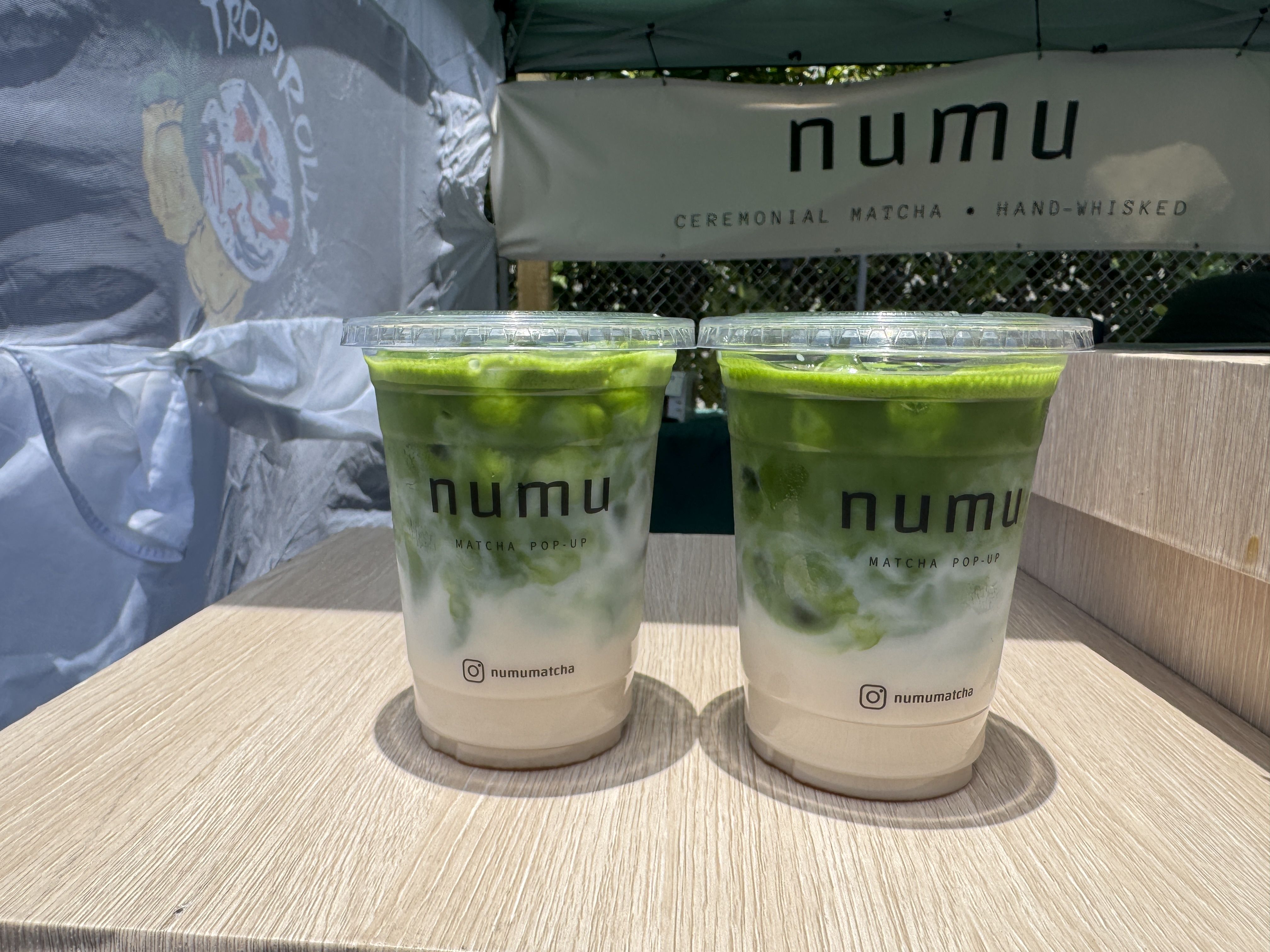 Two iced matcha drinks in clear cups with lids sit on a wooden surface, green swirls visible through the cups. A banner behind shows the brand name numu and the words CEREMONIAL MATCHA.