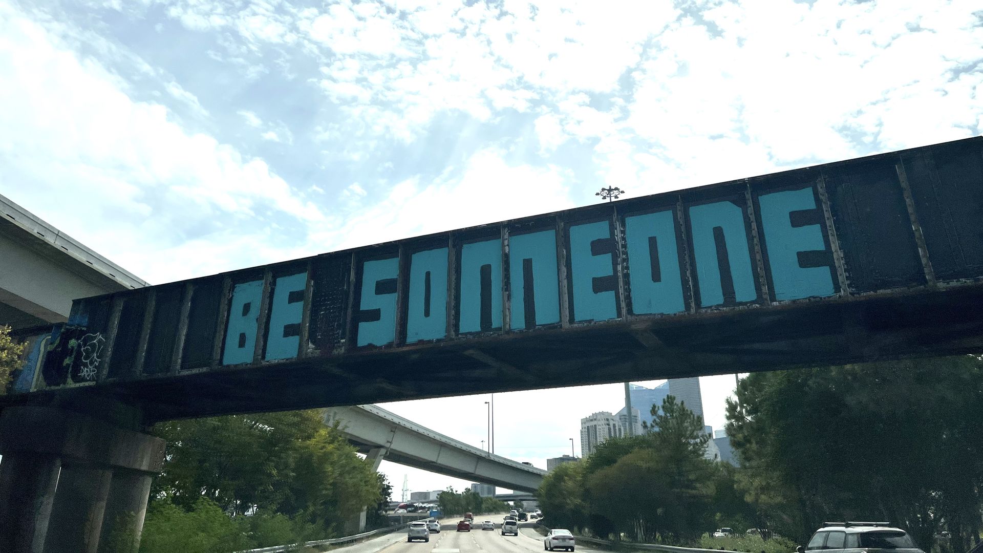The words "Be Someone" inscribed in blue on a train bridge over the freeway