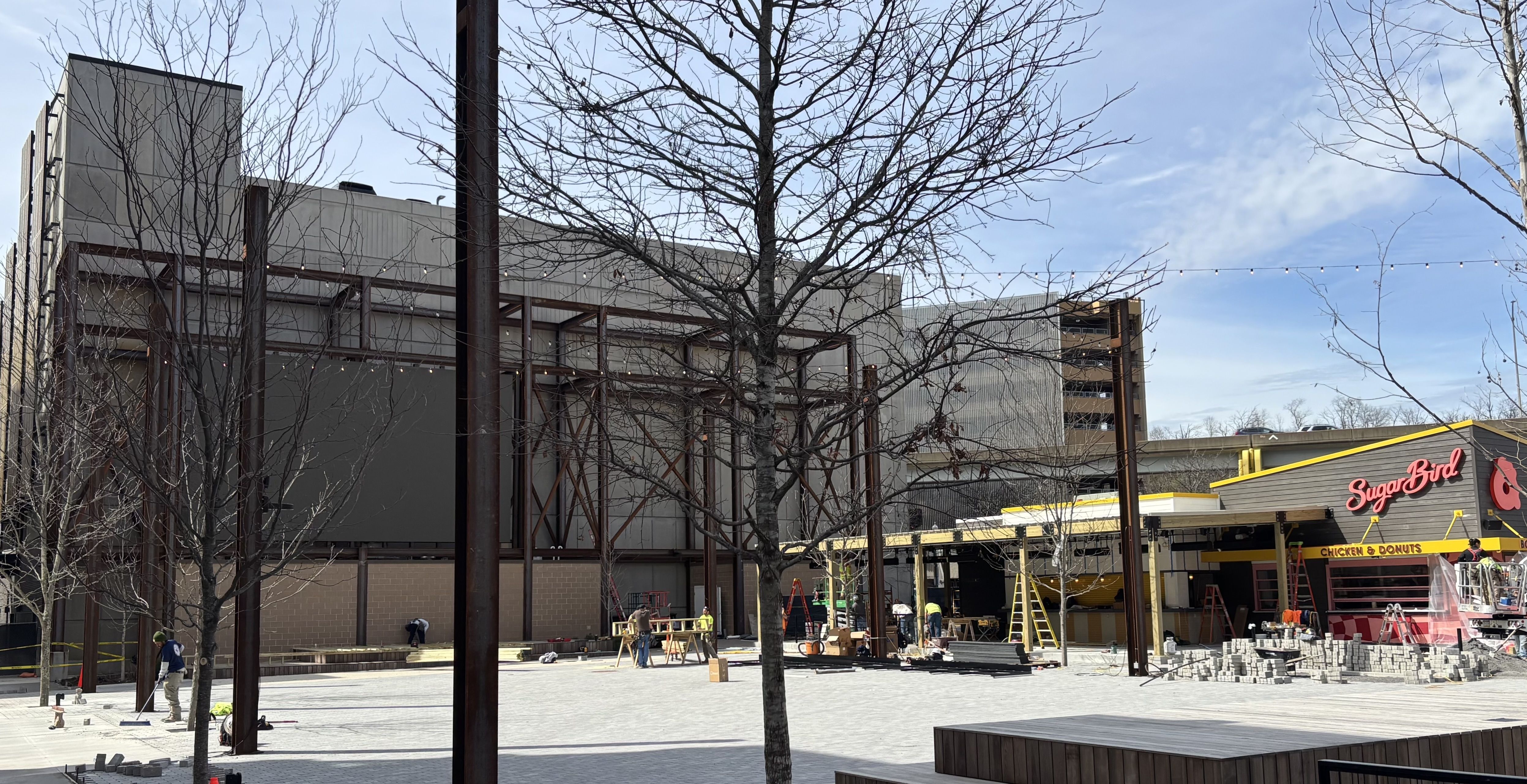 A construction site in a plaza with leafless trees, steel frames, ladders, and workers, near a Sugar Bird restaurant with a yellow and red sign reading "Chicken & Donuts" under a blue sky.
