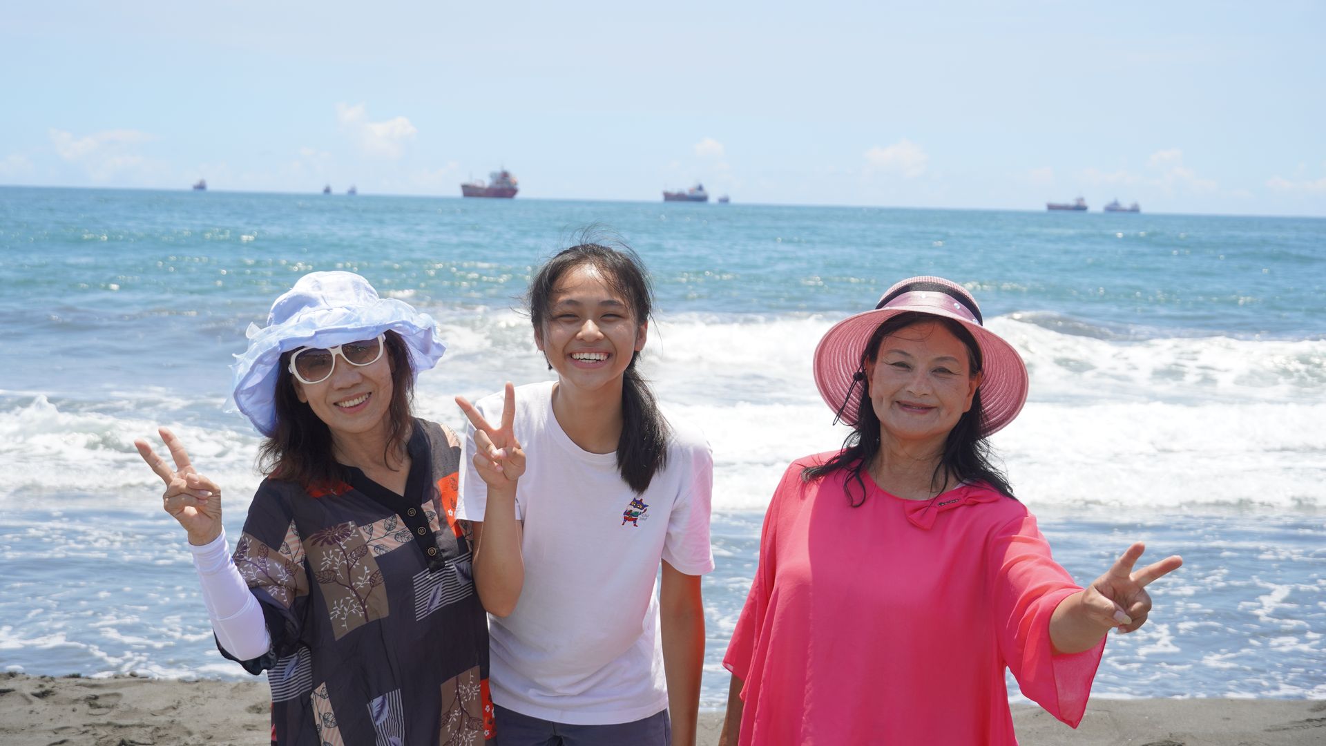 Three tourists pose for a photo on the beach near Kaohsiung Harbor, with oceanliners on the horizon.