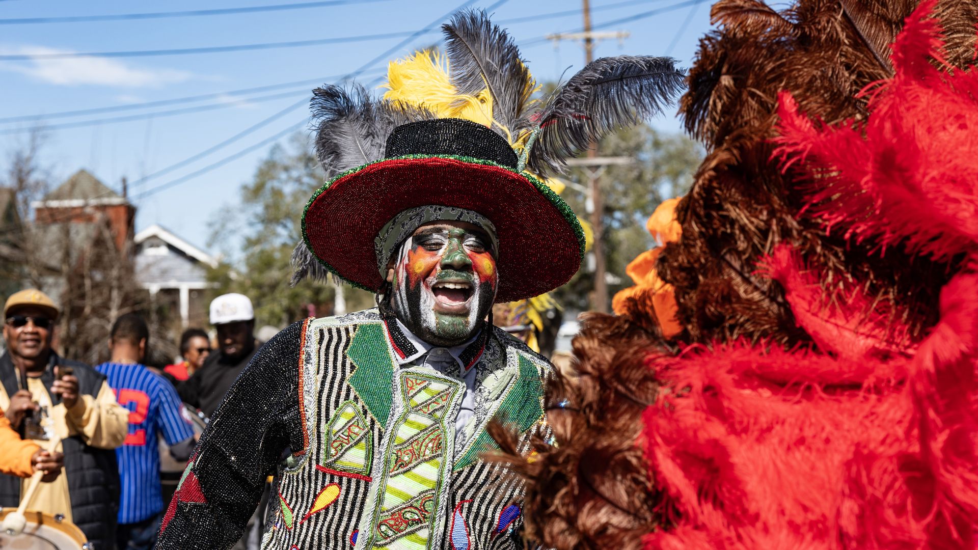 A photo of a Black Masking Indian as he walks in a street next to large red plumage. 