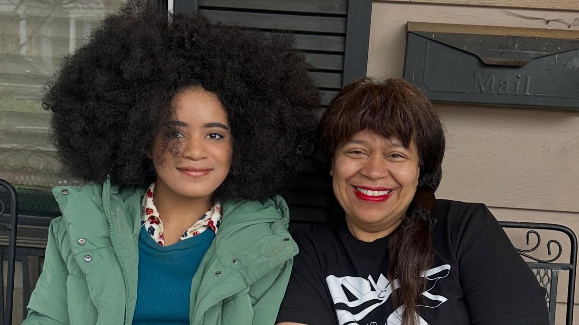 Two women sit on a bench outside a house with a mailbox. Left: a young woman with a large afro, wearing a green jacket and teal top. Right: an older woman with brown hair, black shirt, red lipstick, smiling.