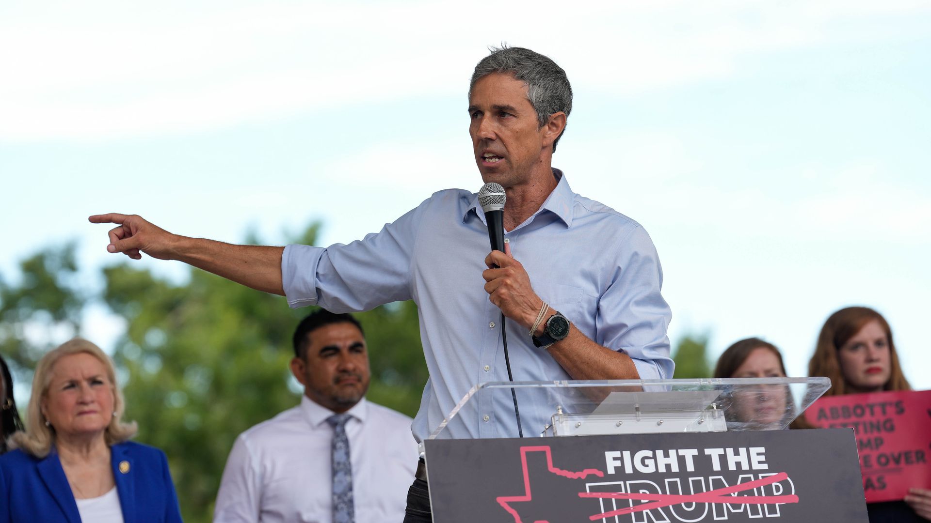 Democrat Beto O'Rourke, wearing alight blue shirt, speaking at a podium with "FIGHT THE TRUMP" sign, pointing his finger. Several people stand behind him, including a woman in blue and a man in a tie, outdoors with trees.