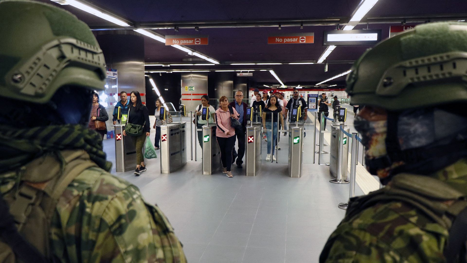 The backs of the heads of two Ecudorian security forces are shown while people are shown walking past gates in a subway in Quito. 