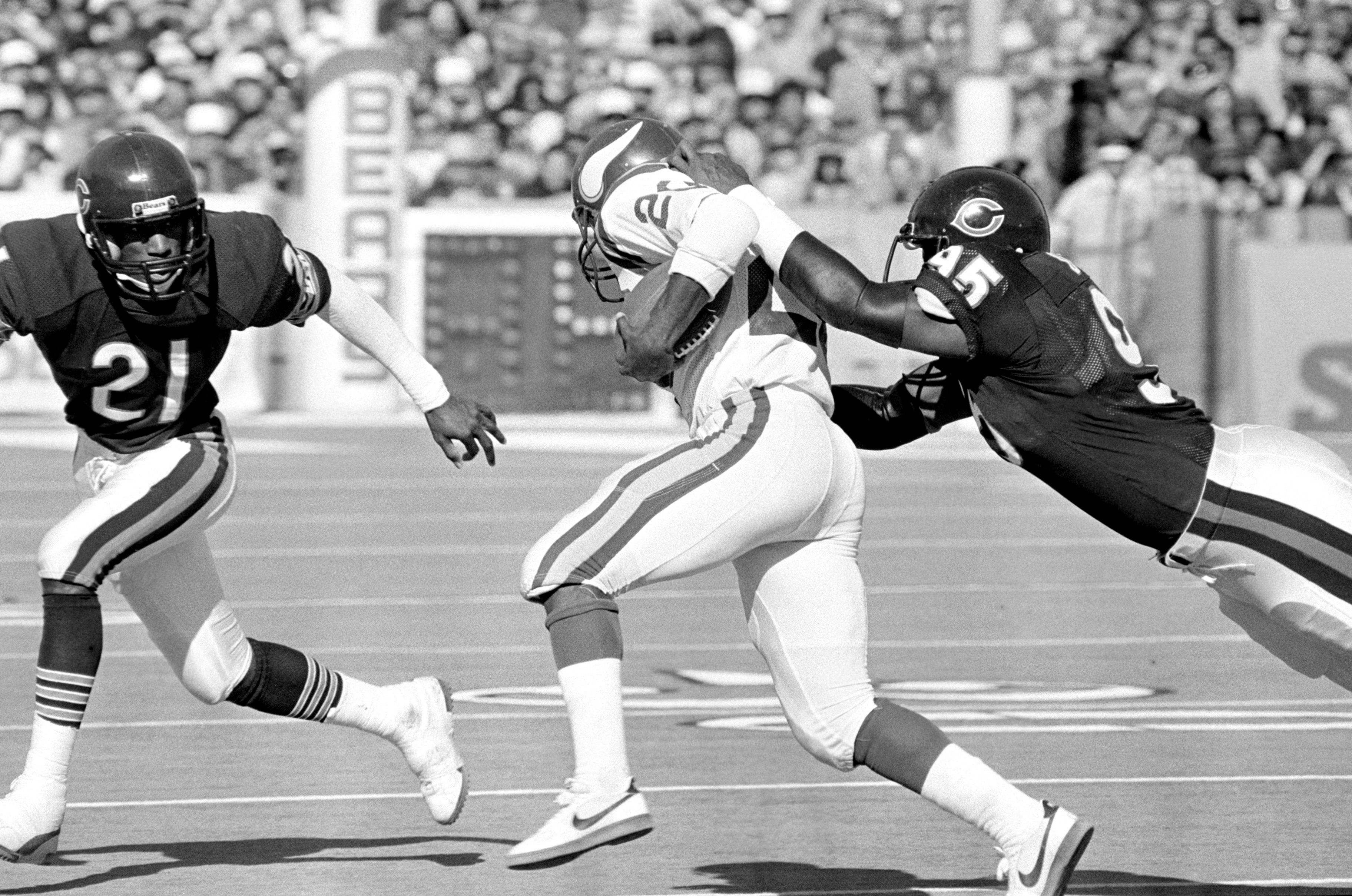 Black and white photo of a football player in light uniform running with the ball while being tackled by a defender wearing number 45 in a dark uniform; another defender approaches.