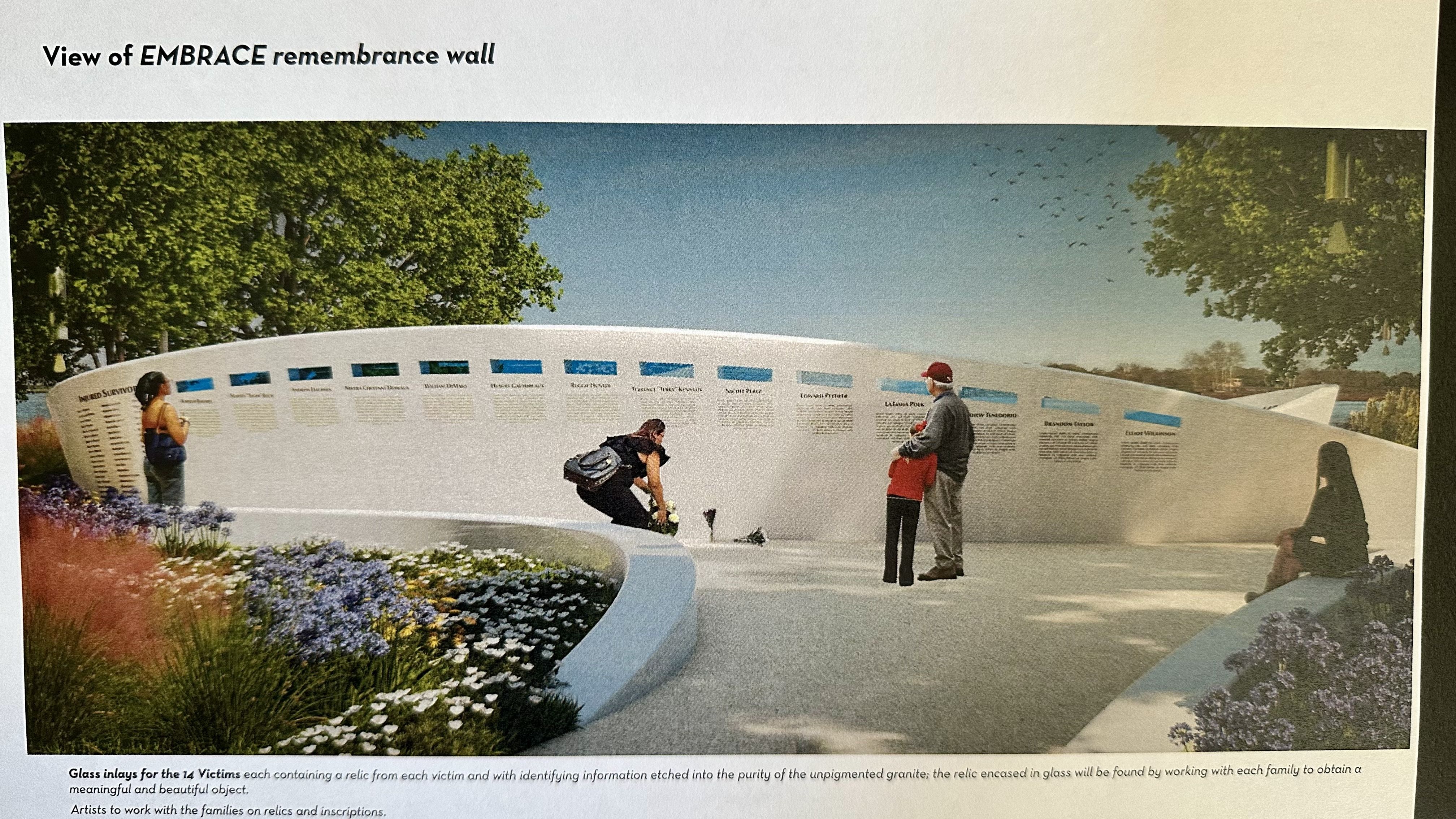 View of EMBRACE remembrance wall: a curved white memorial with blue plaques, visitors reading inscriptions; green trees, clear blue sky, and a flower bed in the foreground.