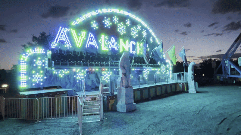 An amusement ride named "Avalanche" glowing with a green and blue neon arch, flashing lights, white railings and a statue at the platform, under a dusky evening sky.