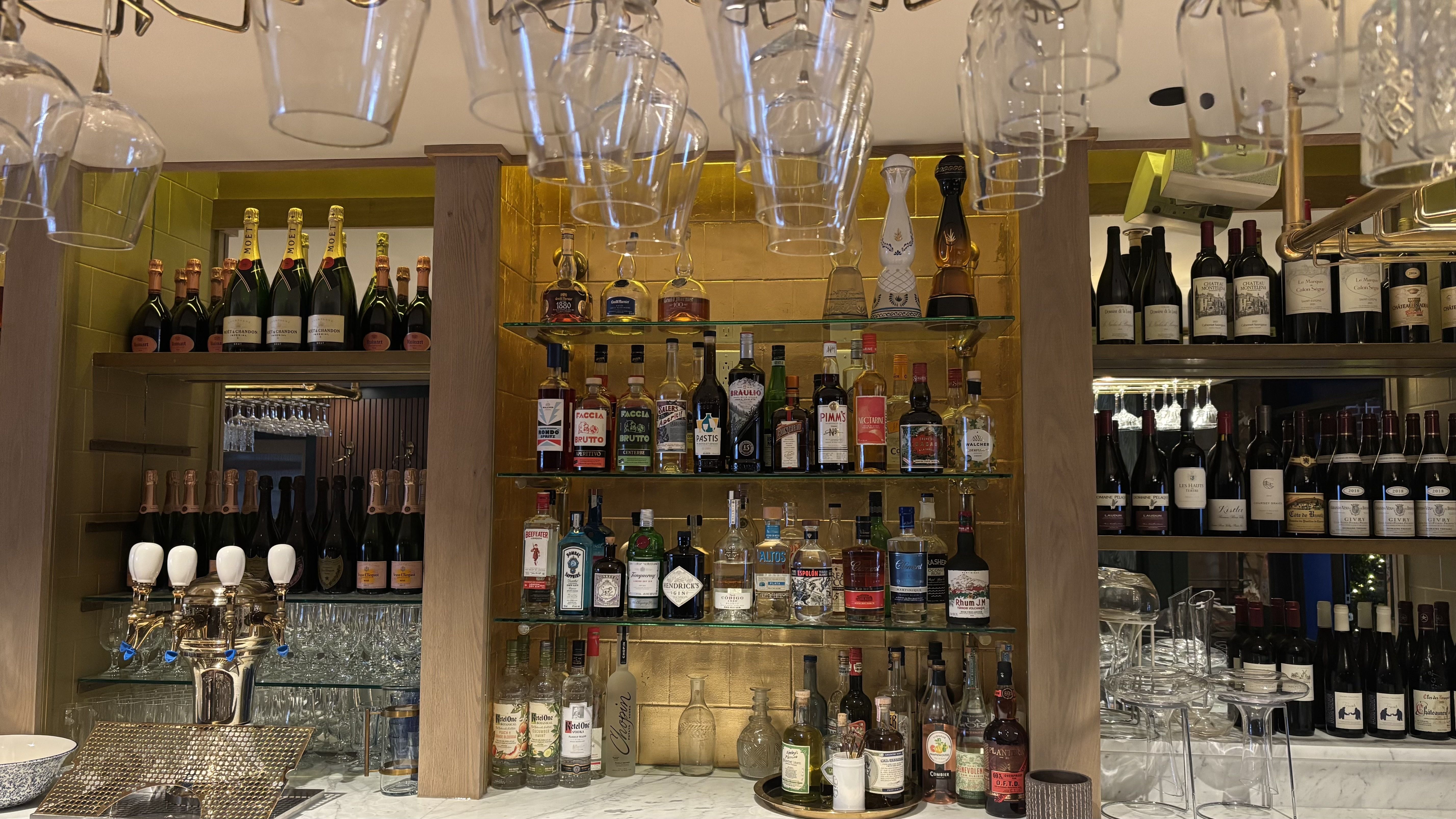 Bar shelf with various liquors, wines, and champagnes, with hanging wine glasses above and glassware below, framed by wooden posts and gold tile back wall.