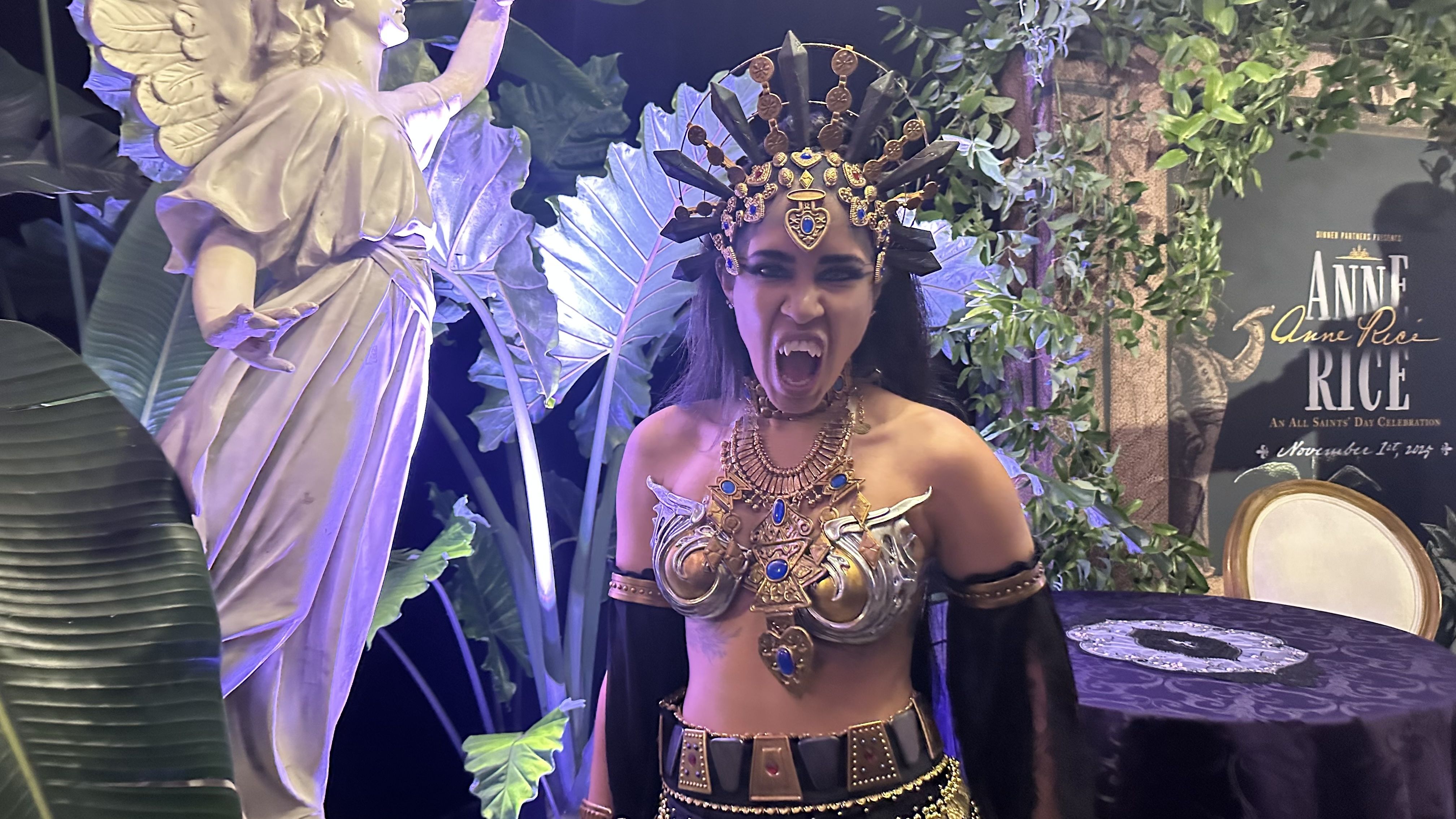 Person in intricate warrior costume with metal and blue gem decorations, black gloves, and headpiece, making a fierce expression with bared fangs, beside a statue and plants.