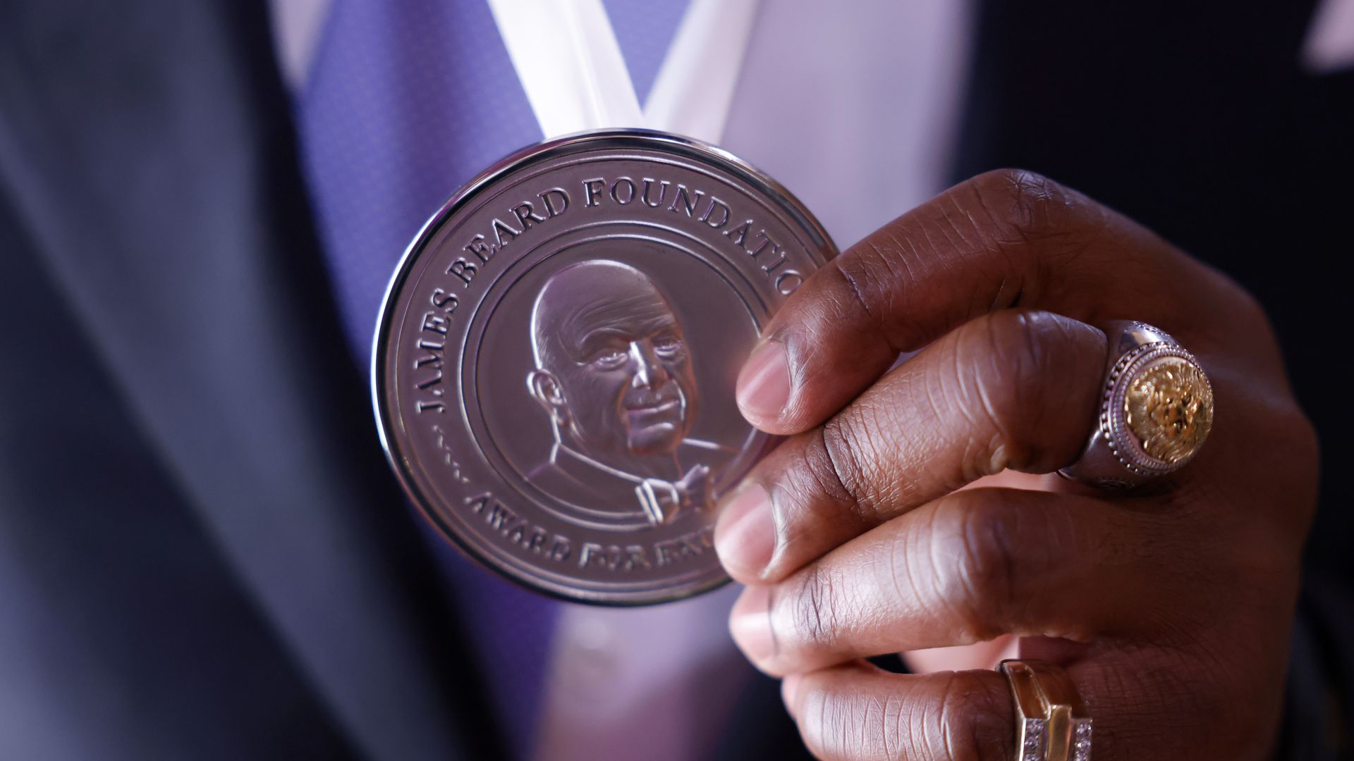 Close-up of a hand holding a large silver James Beard Foundation medal with a portrait; the hand wears a gold signet ring and a diamond-studded ring, with a purple tie and suit in the background.