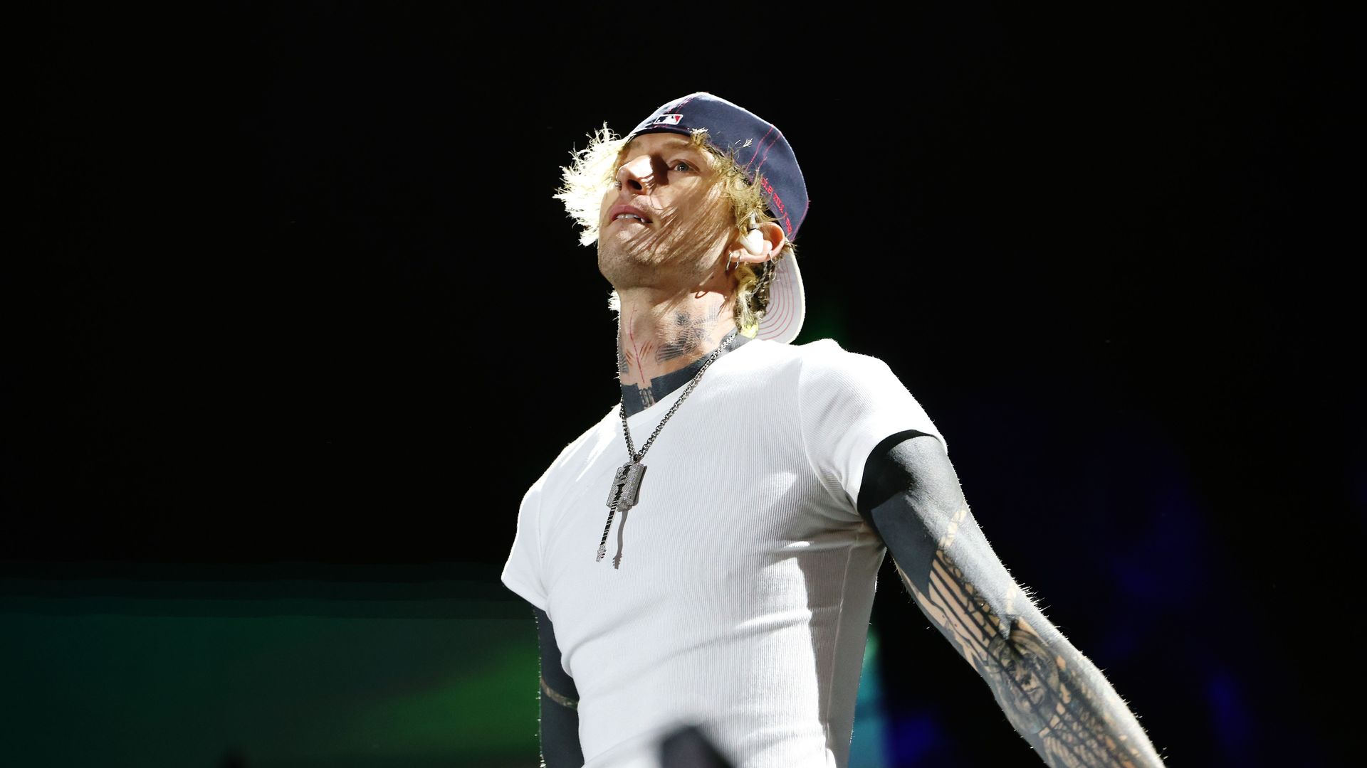 Male performer with a backward baseball cap, white t-shirt over a black long sleeve shirt, extensive tattoos on neck and arms, illuminated on stage against a dark background.
