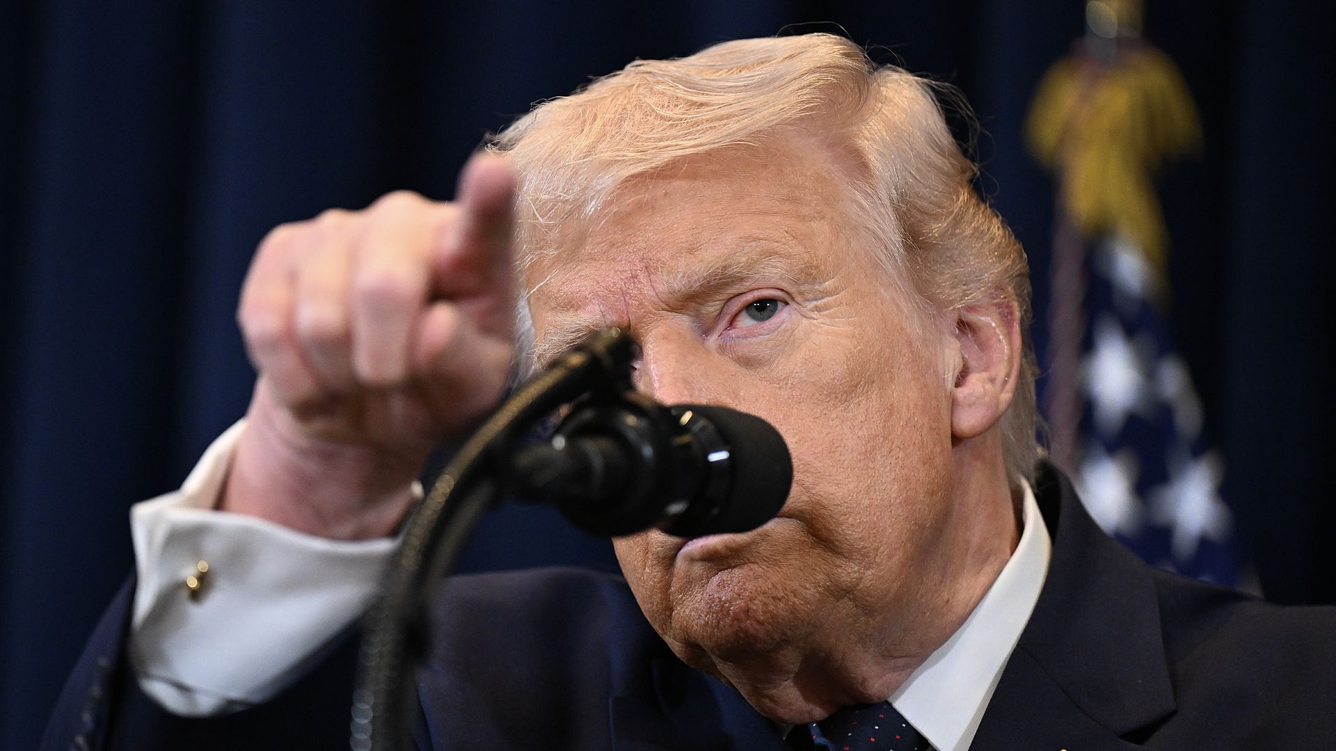 President Trump points during a press conference as he stands behind a microphone while wearing a dark suit, a white collared shirt an American flag pin and a blue tie with red and white spots.