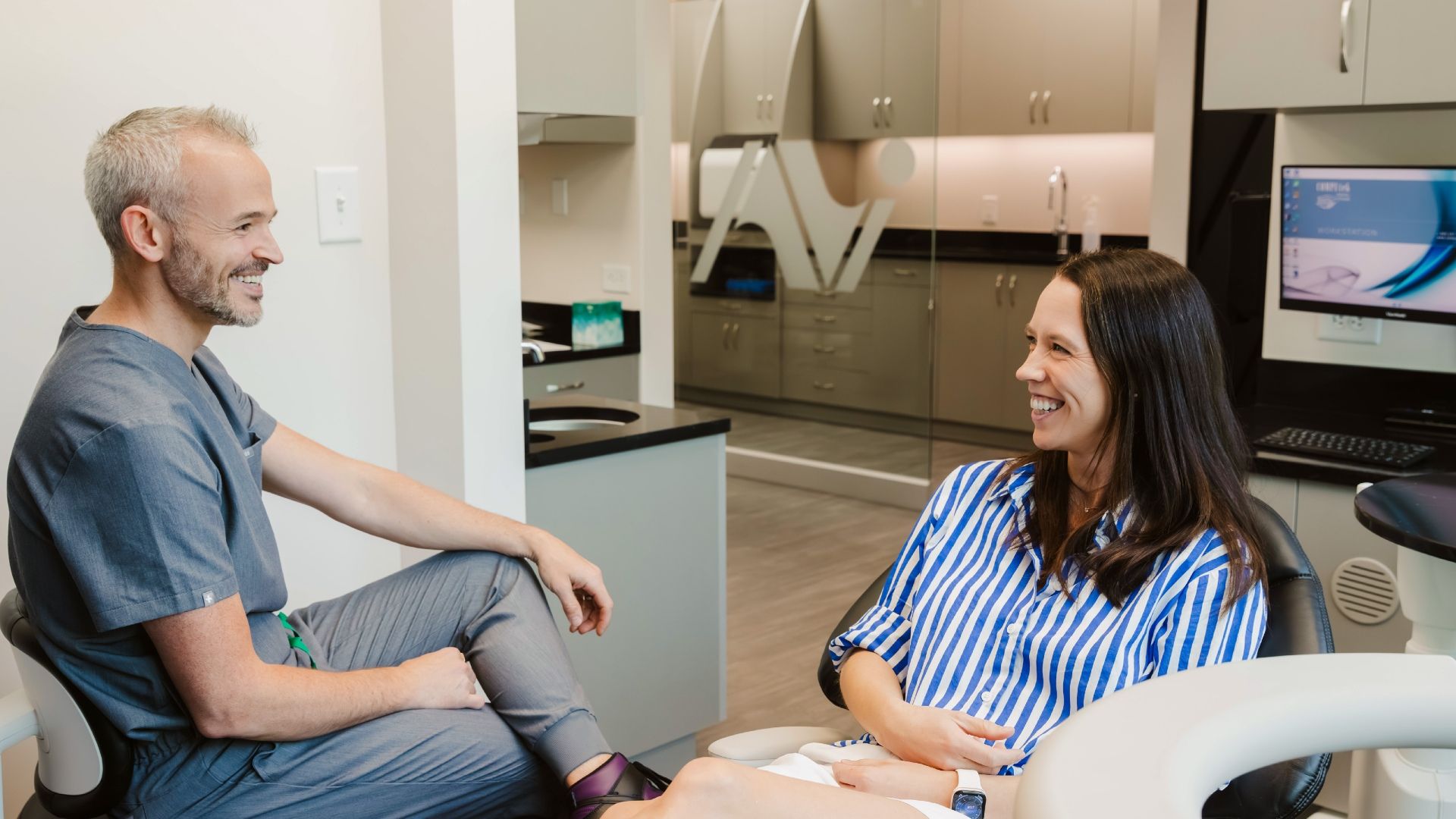 A smiling male clinician in gray scrubs sits across from a smiling female patient in a blue-striped shirt in a modern clinic. Glass partition, cabinets, and a computer monitor are in the background.