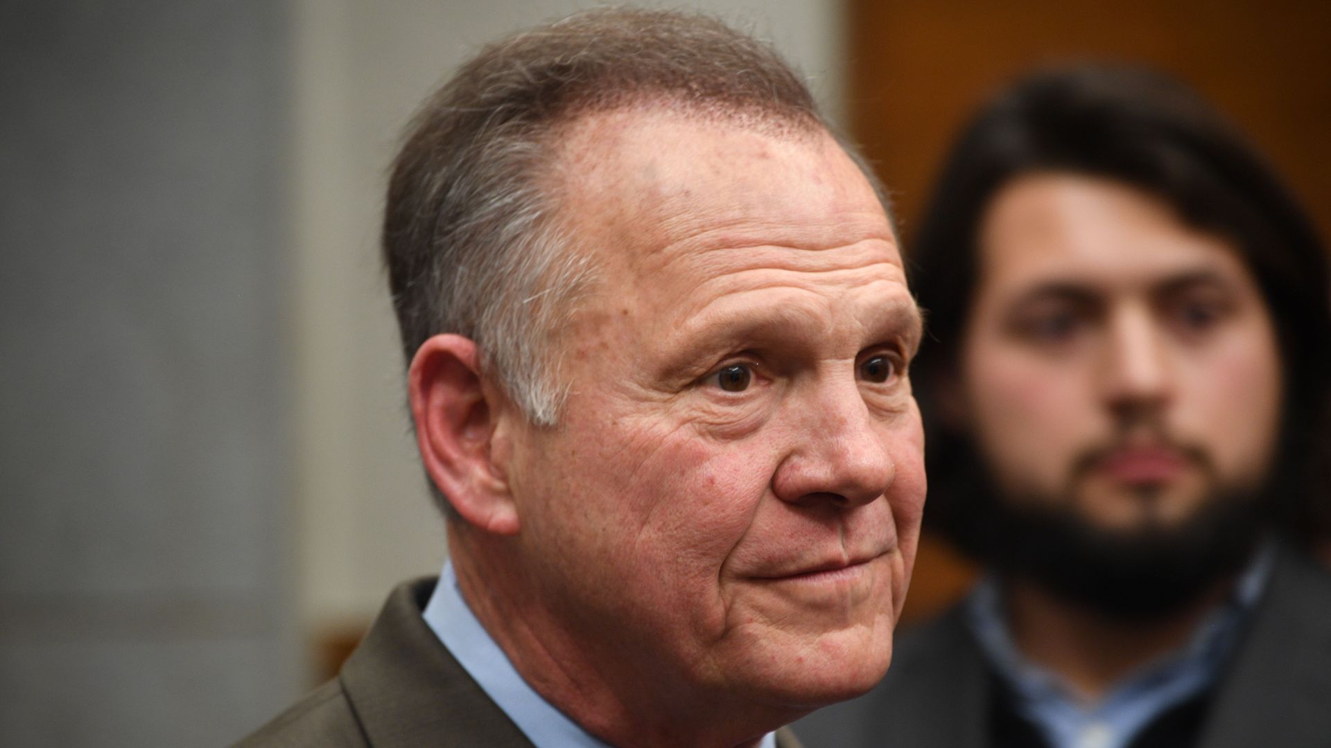 In this image, Roy Moore stands and listens while facing the right.