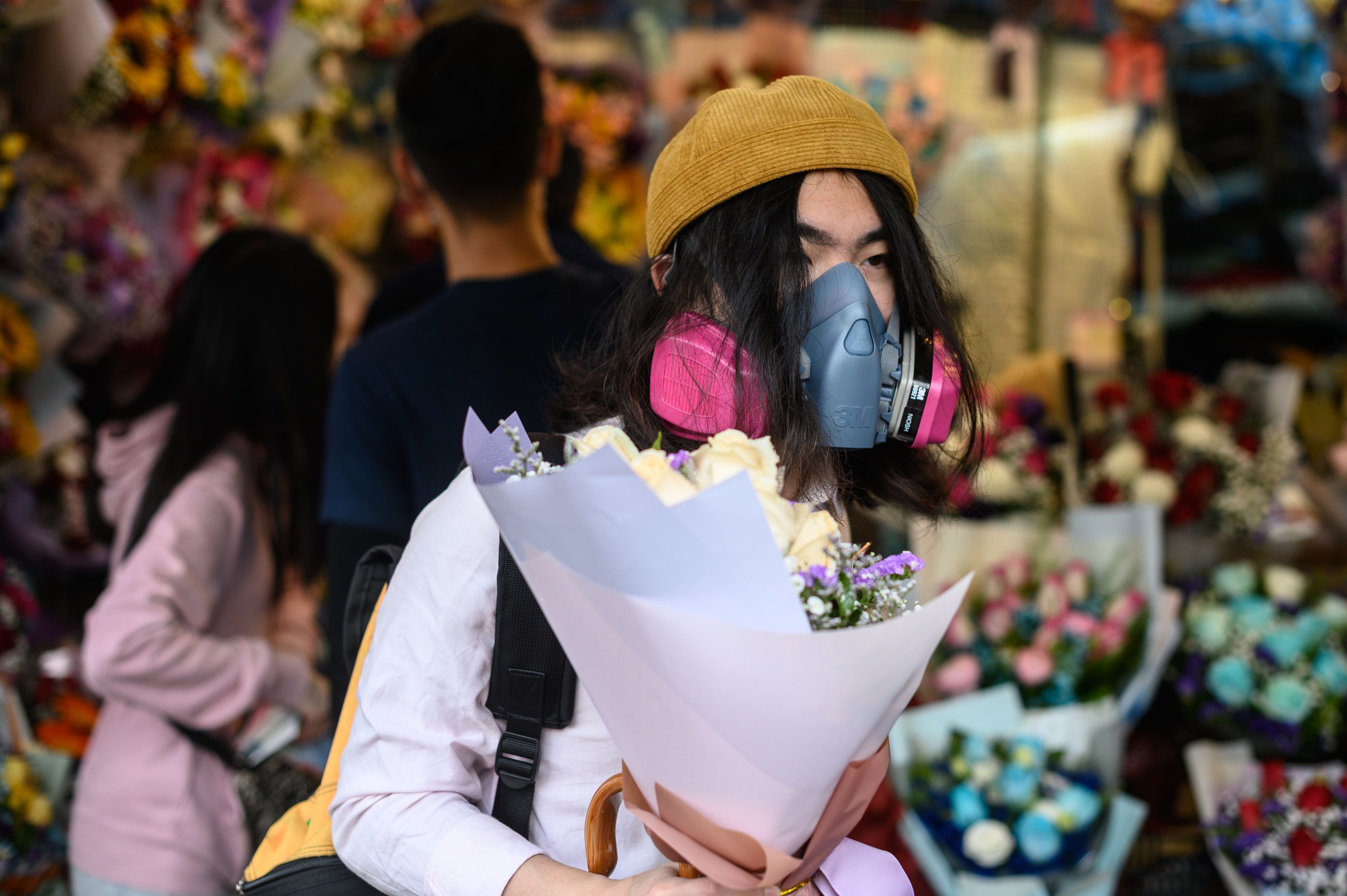 In this image, a woman carries a bouquet of flowers while wearing an industrial gas mask