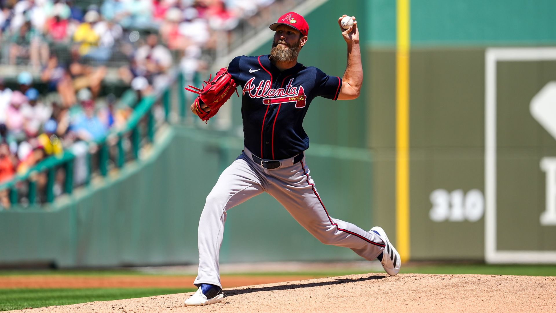 A bearded man wearing a red hat, red baseball glove, blue Atlanta Braves jersey and grey pants throws a baseball as fans seated in green seats watch.