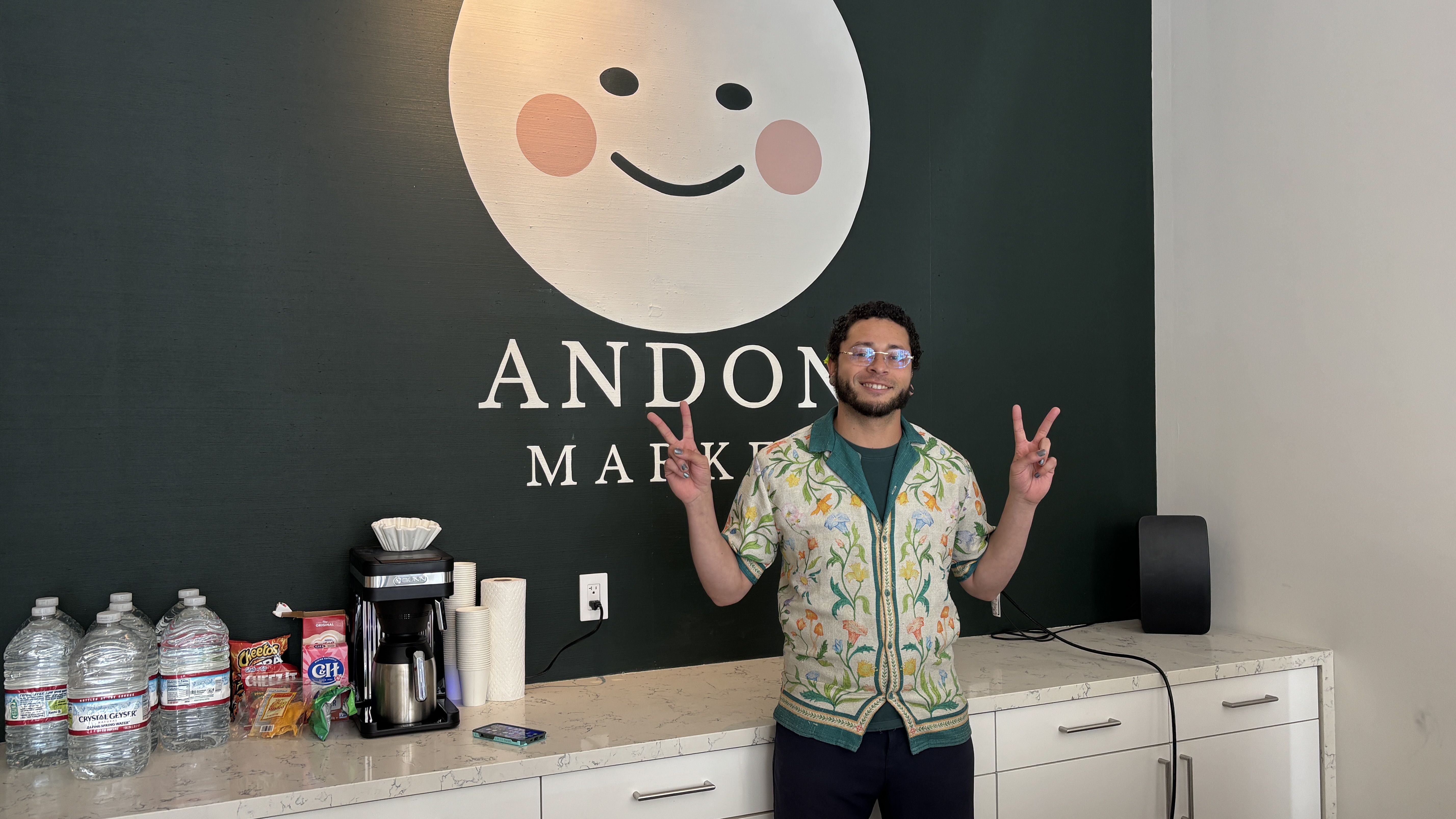 Man with glasses in a floral shirt poses with peace signs in front of a dark green wall featuring a large white smiling circle logo and the text "ANDON MARKET". On the counter: water jugs, a coffee maker, cups, and snacks.