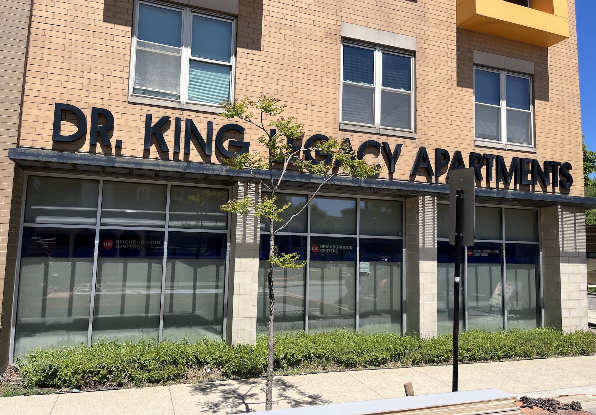 Brick building with large black letters across the facade reading "DR. KING" and "APARTMENTS". A small tree in front, glass storefronts below, and a yellow balcony on the top right against a blue sky.