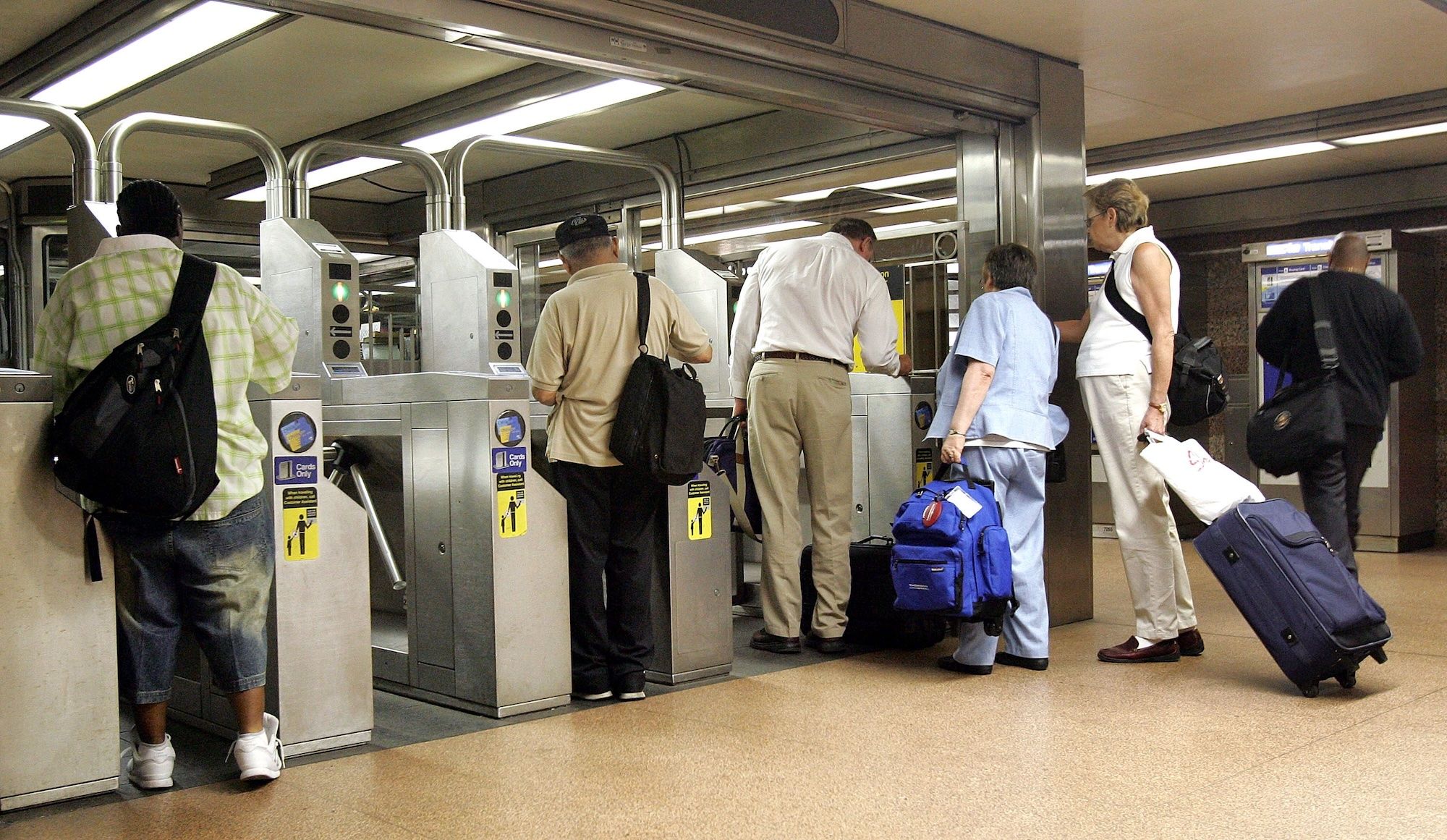 People wait at metal subway turnstiles under bright ceiling lights. A diverse group with backpacks and rolling suitcases lines up to enter, as green indicators glow.