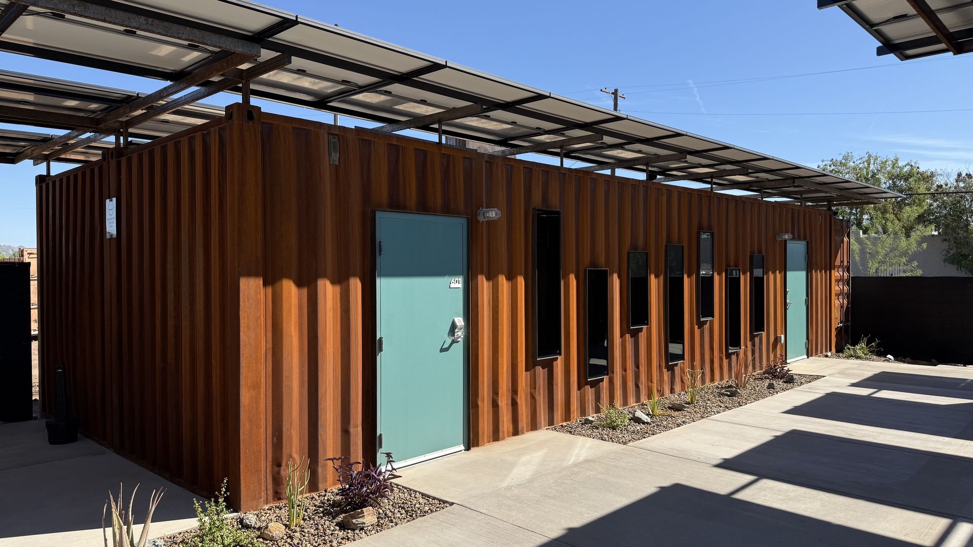 Row of rust-colored shipping containers converted into a building with teal doors and black-framed windows, shaded by a solar-panel canopy; desert landscaping with rocks and drought-tolerant plants.
