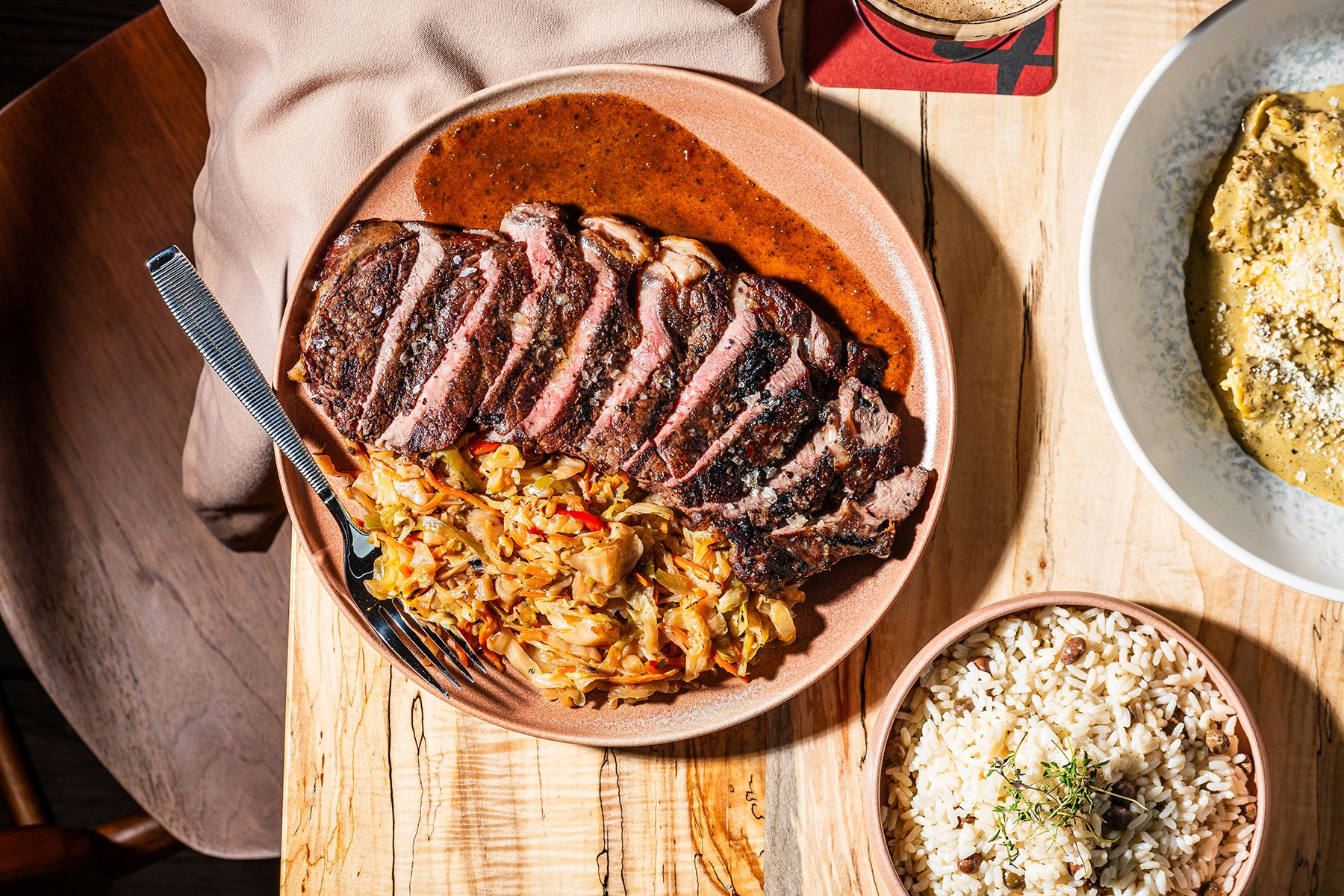 Top-down view of sliced grilled steak on a pink plate with brown sauce, resting on sautéed cabbage and carrots; wooden table, fork, and bowls of rice and a creamy side dish nearby.