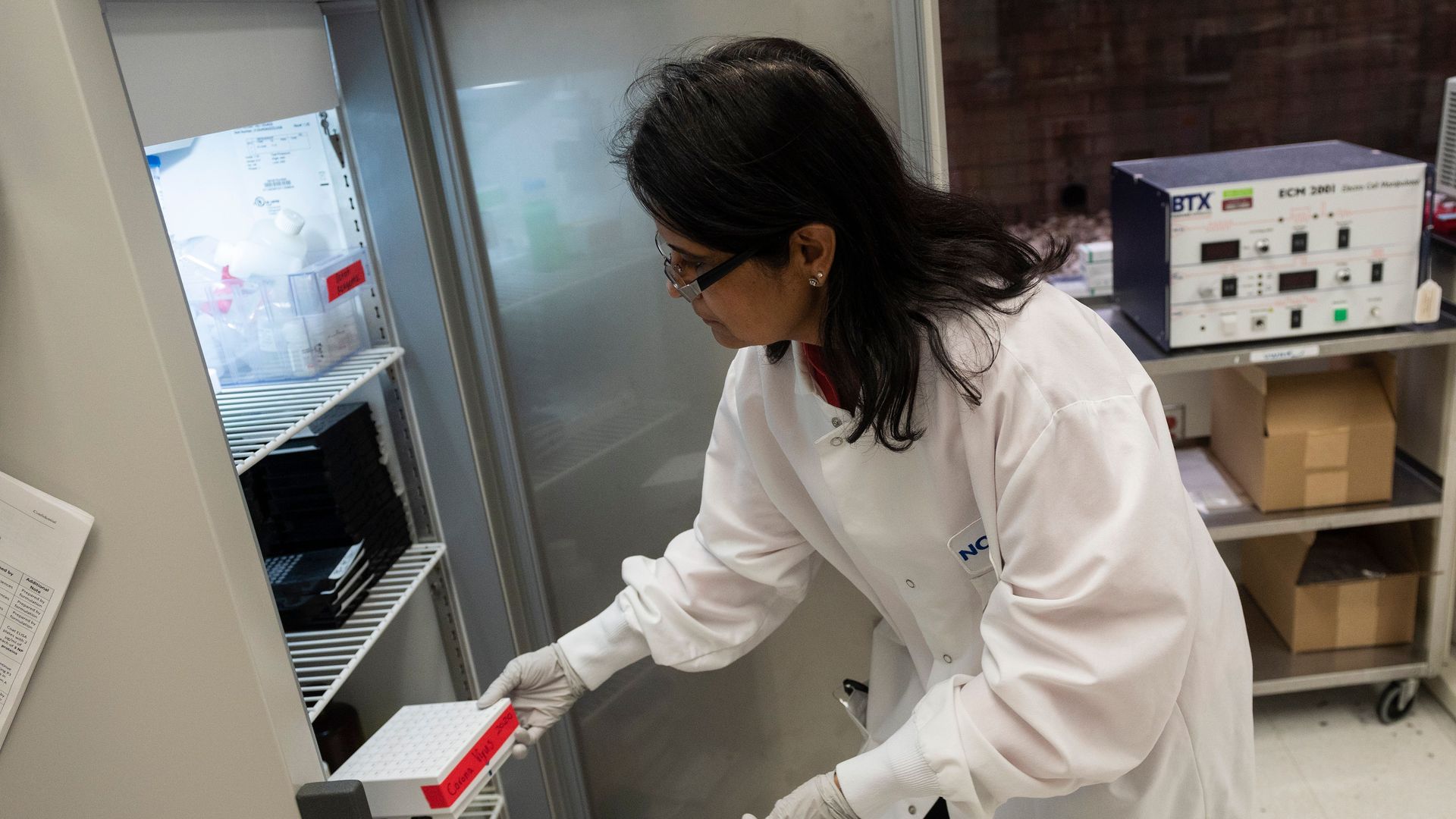 A researcher in a lab coat pulls out a vaccine vial from a fridge.