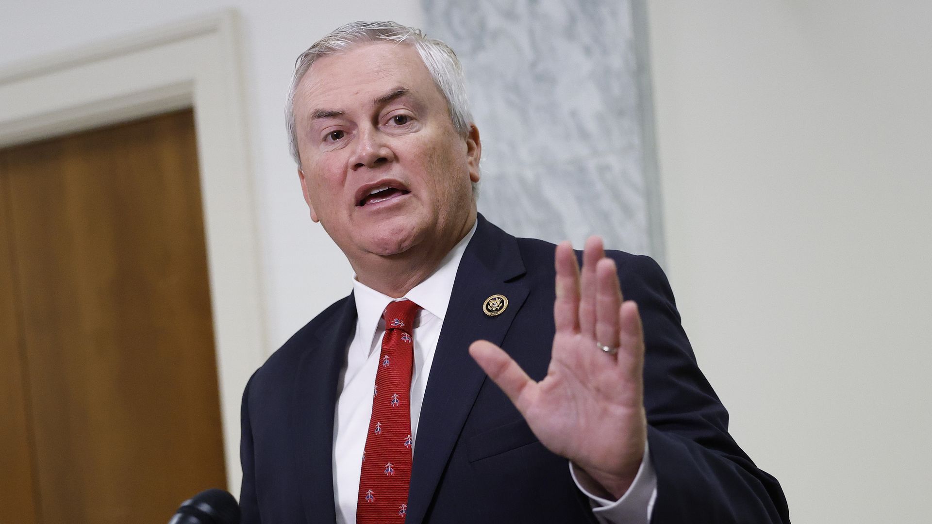 Middle-aged man with gray hair in a dark suit, white shirt, and red tie, speaking and gesturing with his hand raised, standing near a microphone indoors with a wooden door and marble wall behind.