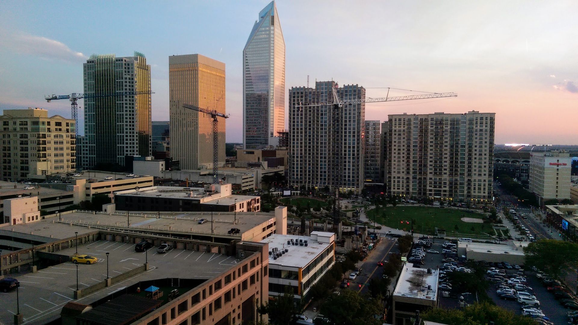 City skyline at dusk with tall buildings, construction cranes, and a park with green lawns. Buildings reflect warm sunset light under a clear sky with soft pastel colors.