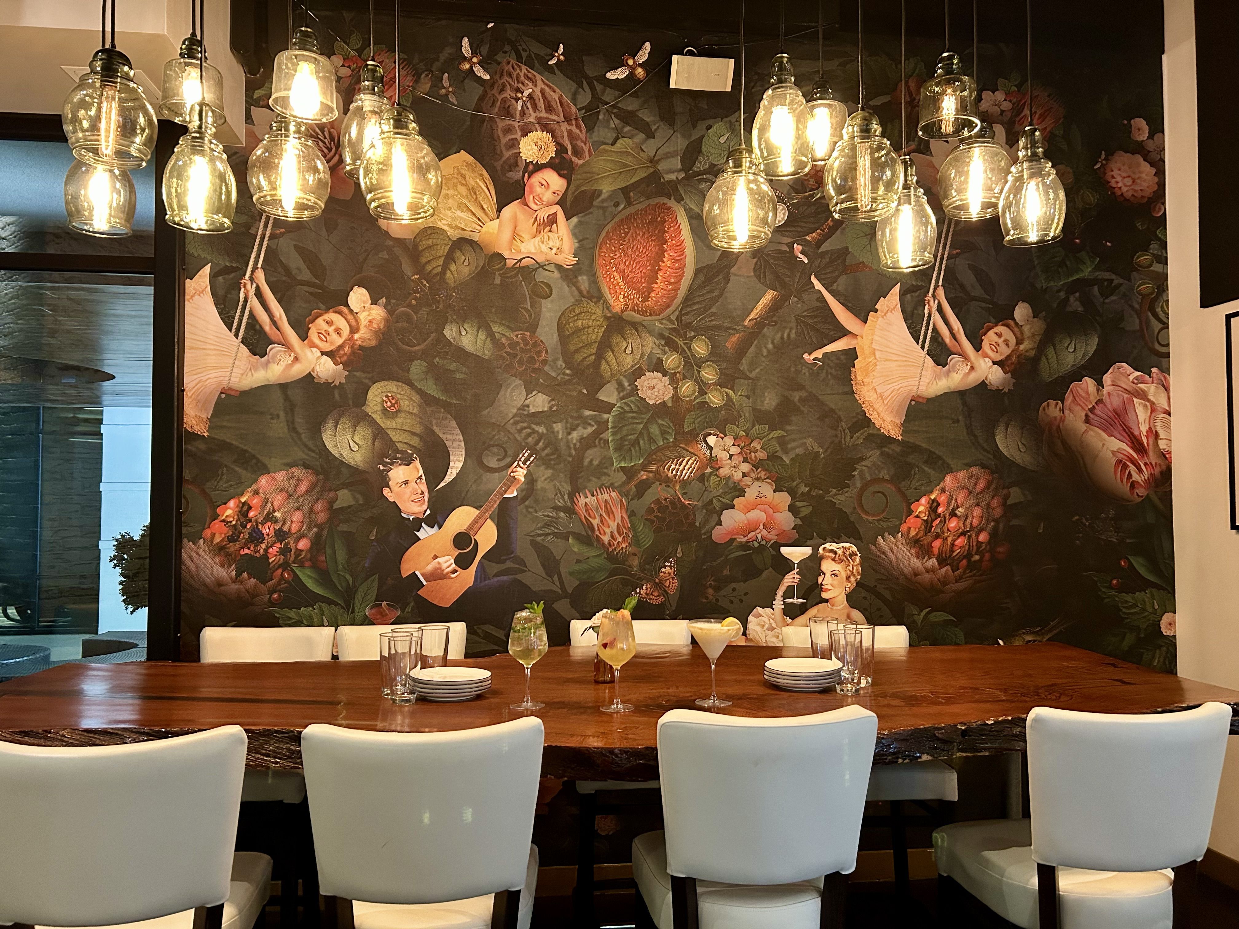 Table with gourmet dishes including flatbread with ham and cheese, a bowl with poached egg and tomatoes, another bowl with bread, and two yellow cocktails with mint garnish in a bright restaurant.