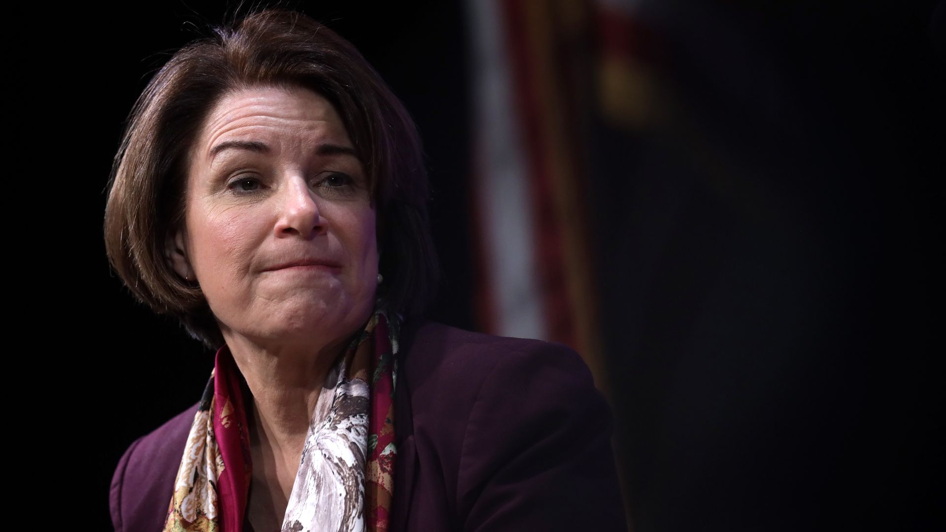 In this picture, Amy Klobuchar listens while sitting and wearing a suit jacket