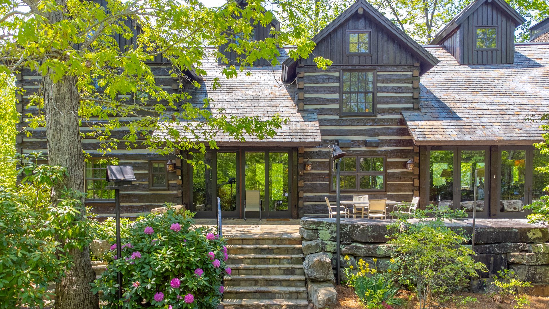 Rustic log cabin with dark wooden beams and a stone staircase leading to a patio with table and chairs, surrounded by green trees and flowering bushes.