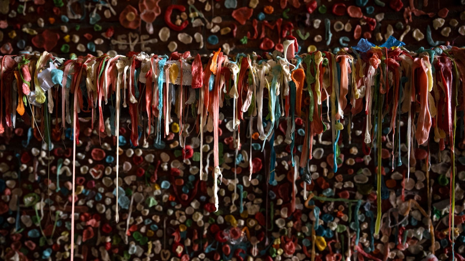 Different colors of chewing gum are stuck to a brick wall, with some stretched to hang long and others stuck in small round wads against the brick.