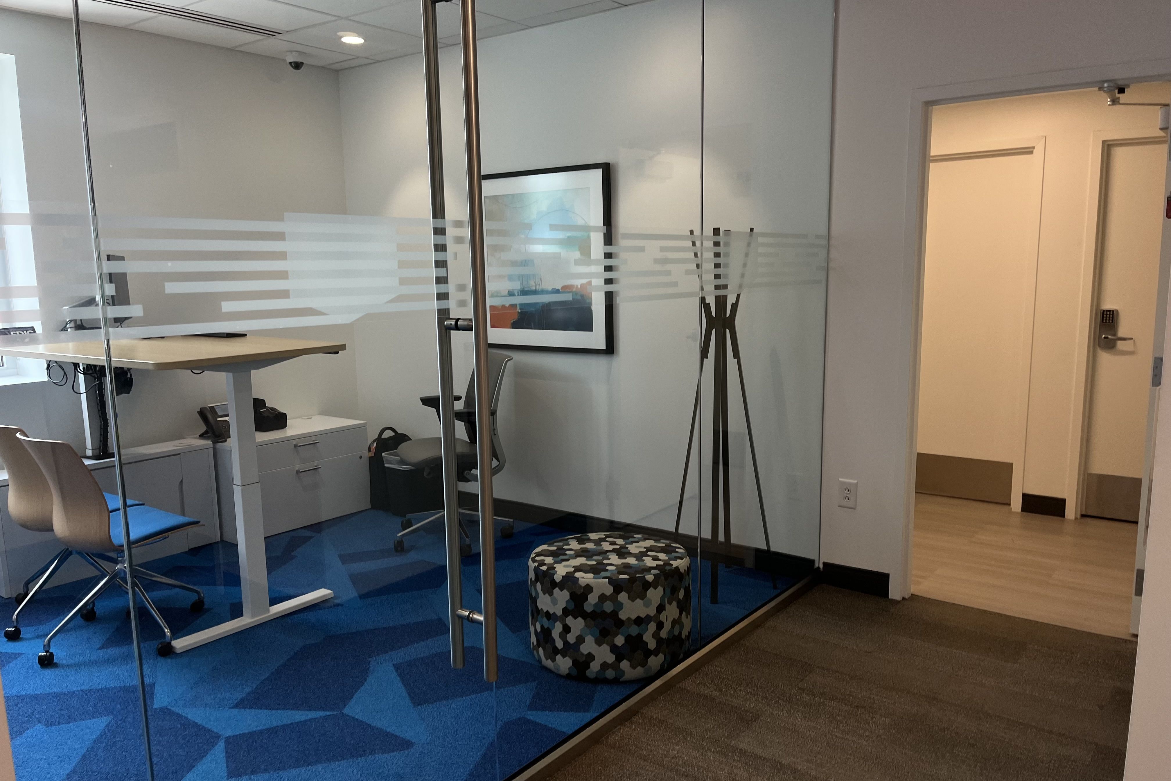 Modern office with glass walls, blue geometric carpet, standing desk, two chairs, round patterned ottoman, coat rack, framed artwork, and hallway with light wood floor.