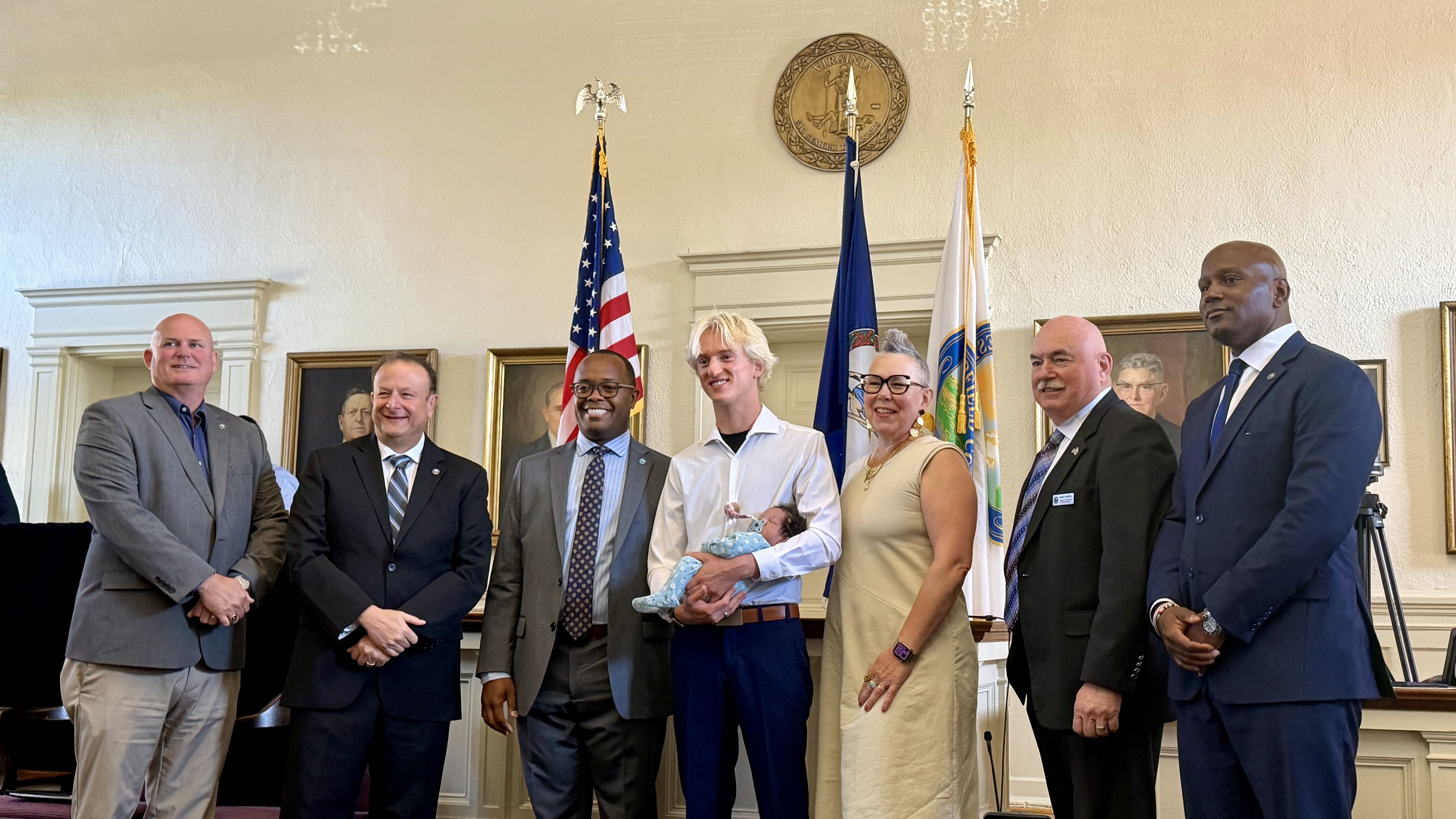Seven adults in business attire pose in a formal room with three flags (American, blue seal, white). A woman in the center holds a swaddled baby; a Virginia seal hangs on the wall behind them.