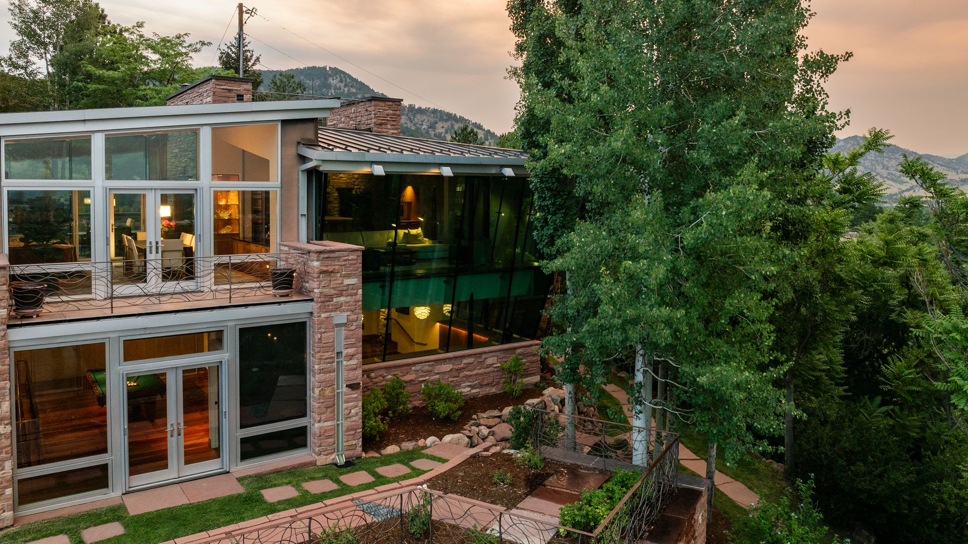 Multi-level house with large glass windows and stone walls, surrounded by green trees and a garden, with mountains in the background.
