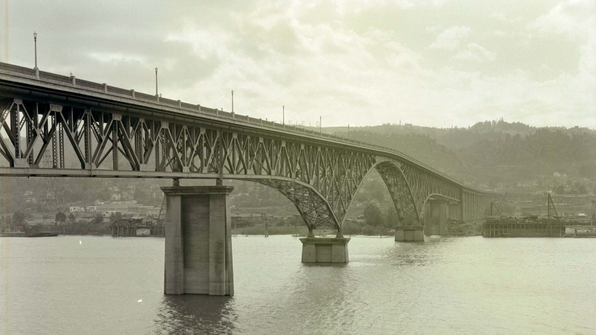 Sepia-toned image of a long steel truss bridge crossing a wide river, supported by tall concrete piers. Distant hills and a small town line the far shore under a cloudy sky.