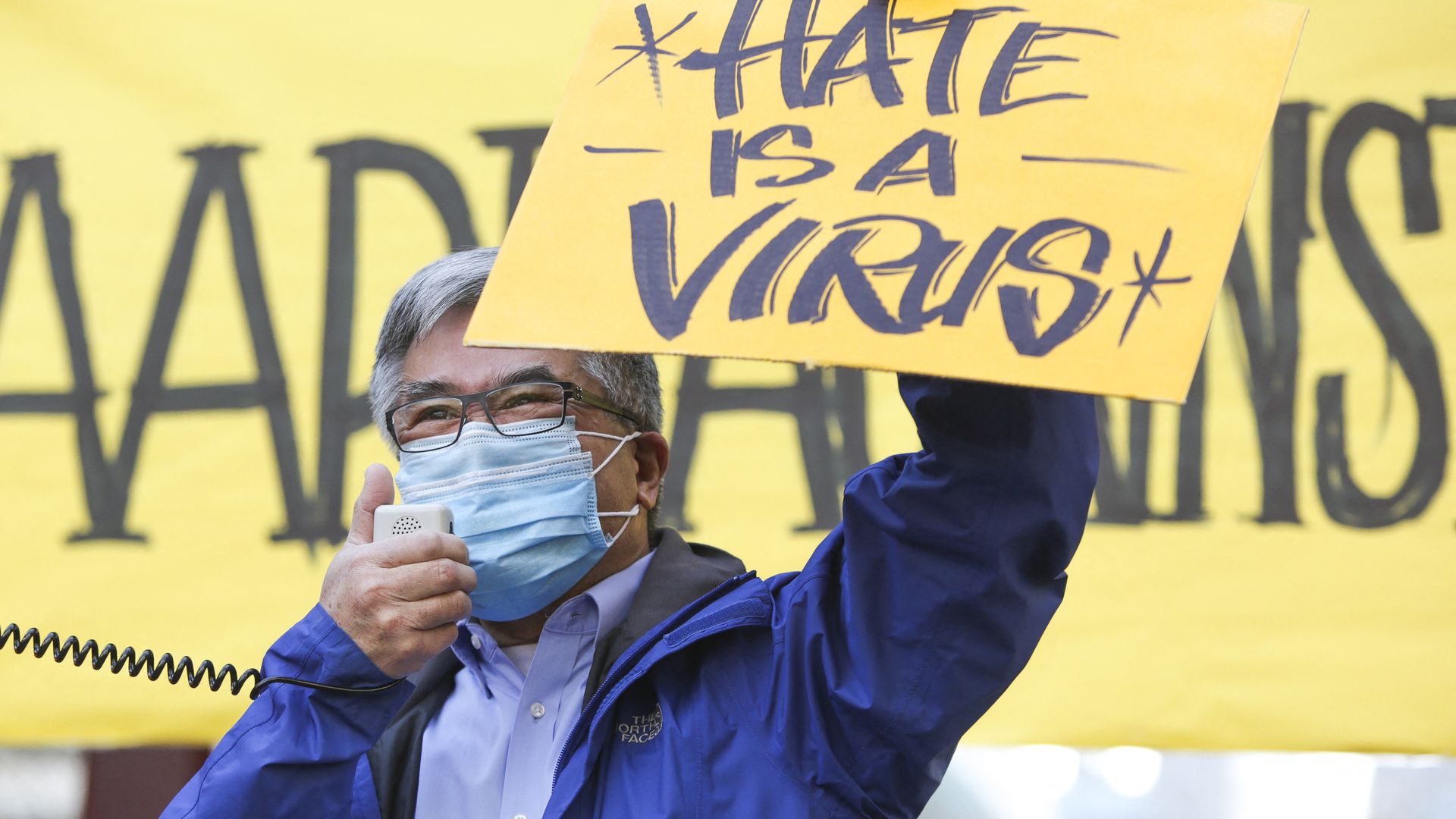 Photo of a masked Gary Locke holding up a yellow sign that says "Hate is a virus"