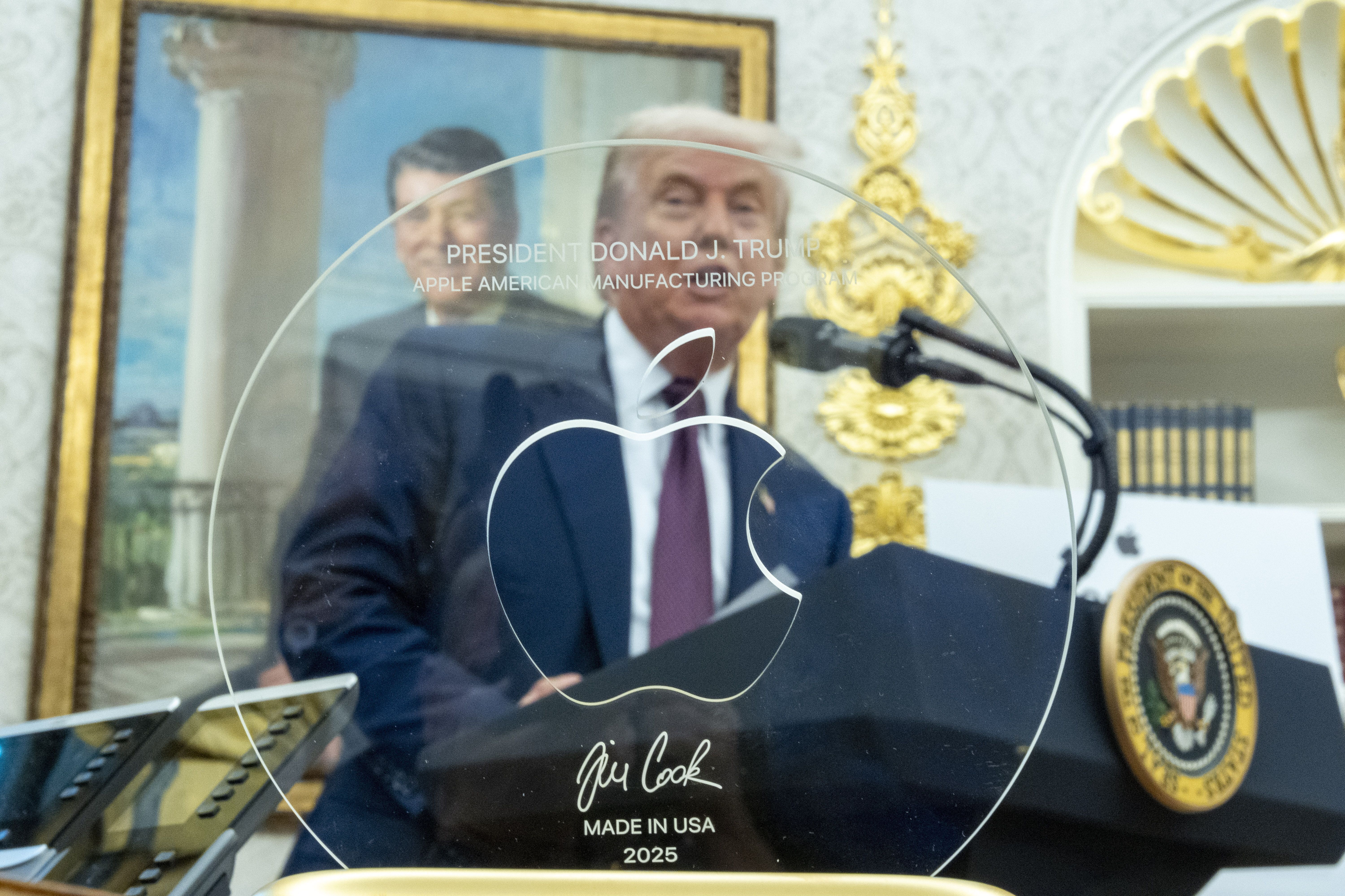 A picture of a clear Apple award plaque with logo and text saying "President Donald J. Trump," "Apple American Manufacturing Program," "Tim Cook," and "Made in USA 2025" with Trump speaking at a podium in the background.
