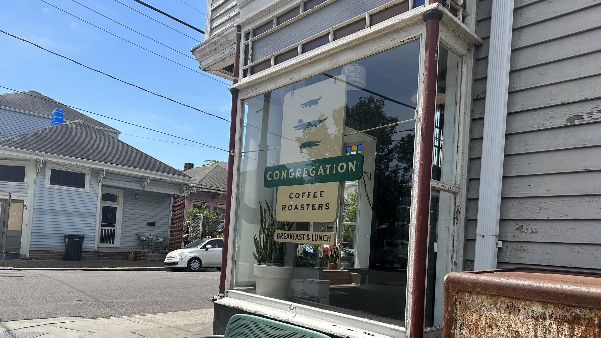 The front window of Congregation Coffee is viewed on a sunny day. The shop's sign, which says Congregation Coffee Roasters: Breakfast and Lunch" and pictures three alligators, hangs in the window.