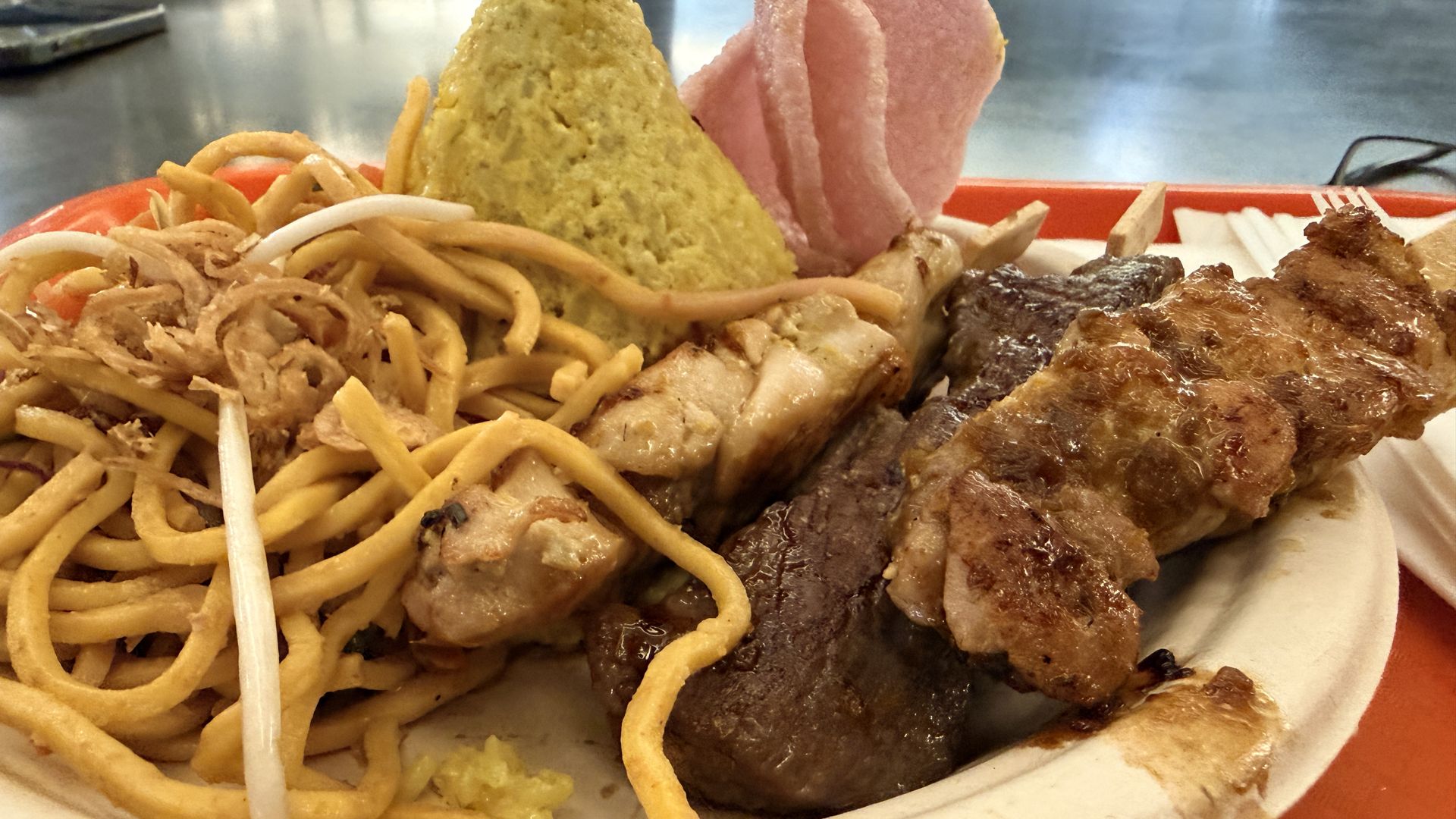 Plate with spaghetti and vegetables on the left, a greenish cone of mashed potatoes in the center, pink ham slices behind, and a large browned steak on the right; stainless diner table in the background.